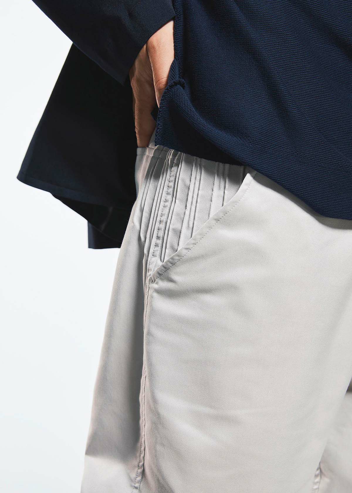 A person wearing TYPE-S BASICS by A-POC ABLE ISSEY MIYAKE—a navy blue top and light beige cropped pants with pleated details, crafted from stretch fabric using the innovative Steam Stretch technique—has one hand tucked into the pants pocket. view 9 of 9