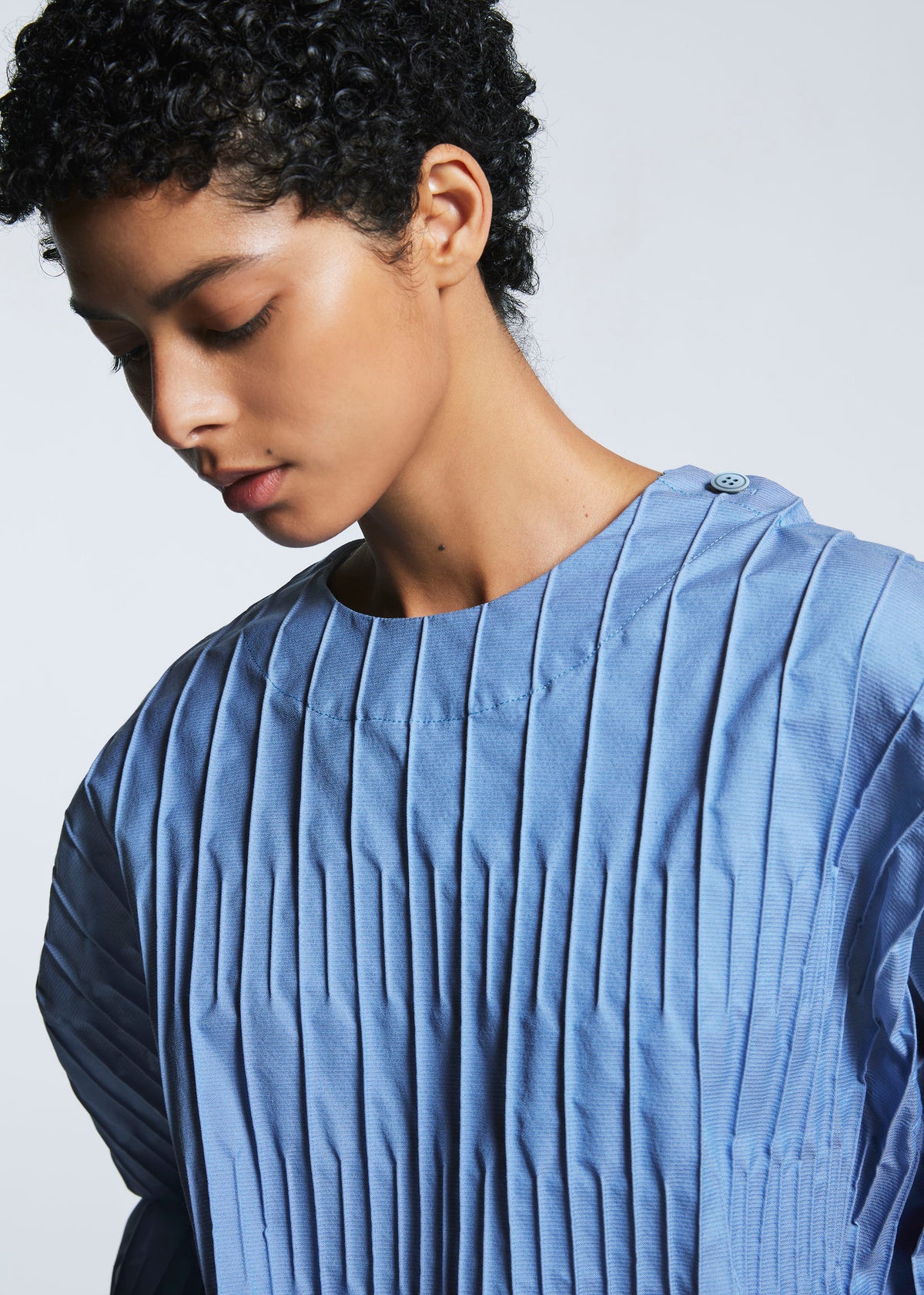A person with short curly hair looks down, wearing the TYPE-W 007 top by A-POC ABLE ISSEY MIYAKE—light blue, round neckline, buttoned shoulder, and loose short sleeves—against a plain background. view 4 of 6