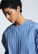 A person with short curly hair looks down, wearing the TYPE-W 007 top by A-POC ABLE ISSEY MIYAKE—light blue, round neckline, buttoned shoulder, and loose short sleeves—against a plain background.