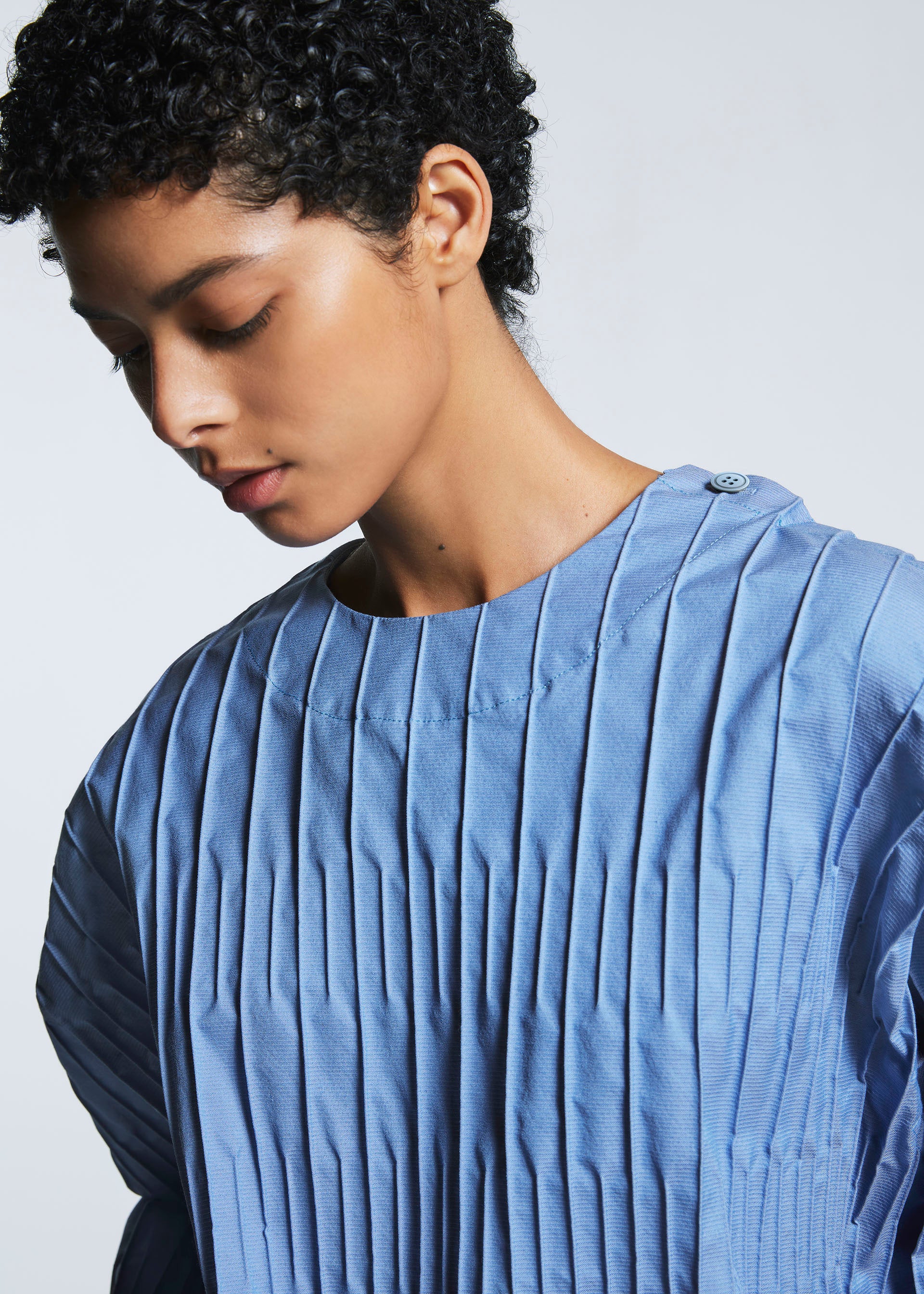 Zoomed view A person with short curly hair looks down, wearing the TYPE-W 007 top by A-POC ABLE ISSEY MIYAKE—light blue, round neckline, buttoned shoulder, and loose short sleeves—against a plain background.