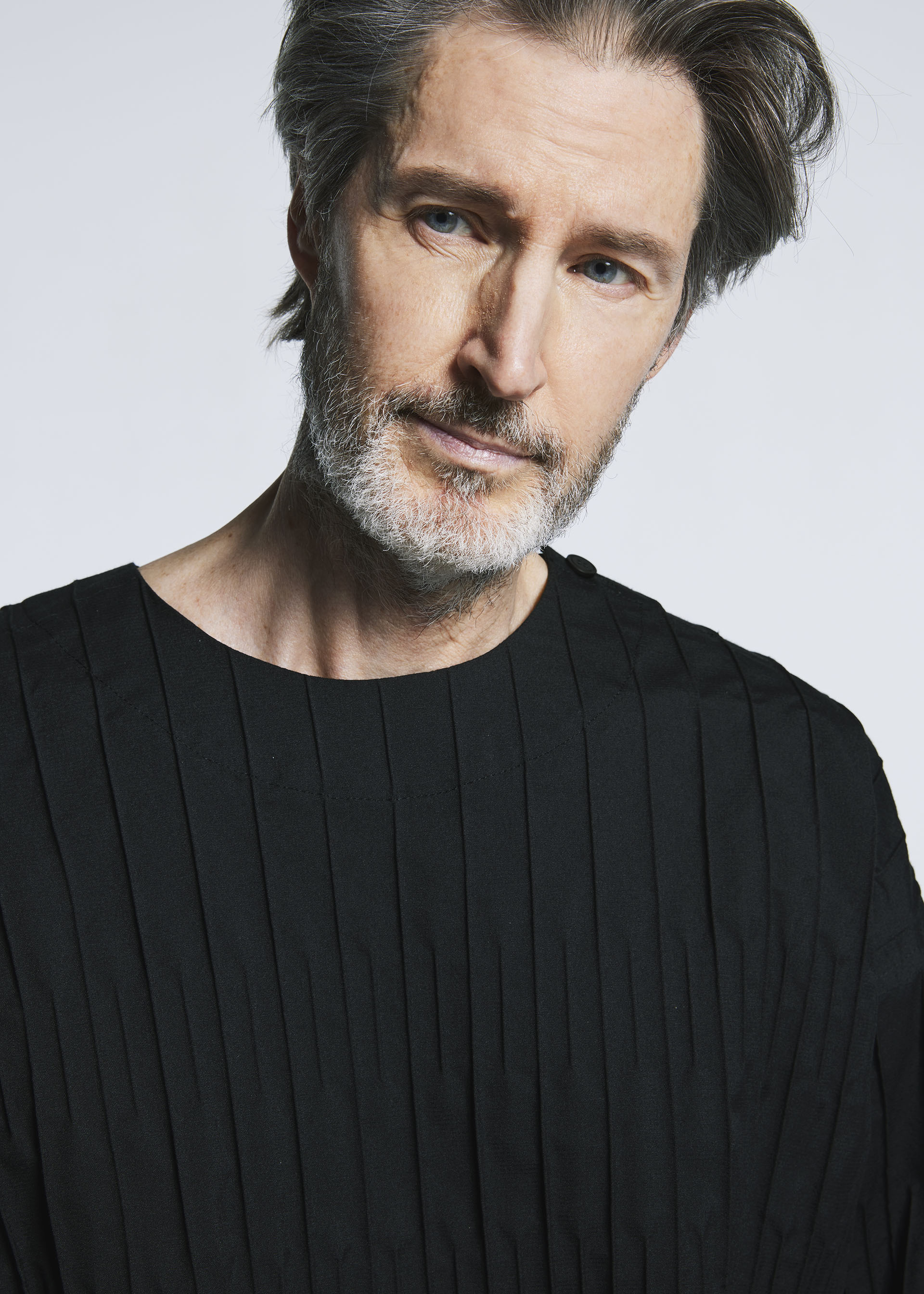 Zoomed view A middle-aged man with gray hair and a beard, wearing the TYPE-W 007 top by A-POC ABLE ISSEY MIYAKE—black with pleated stripes—looks slightly to the side against a plain light background.