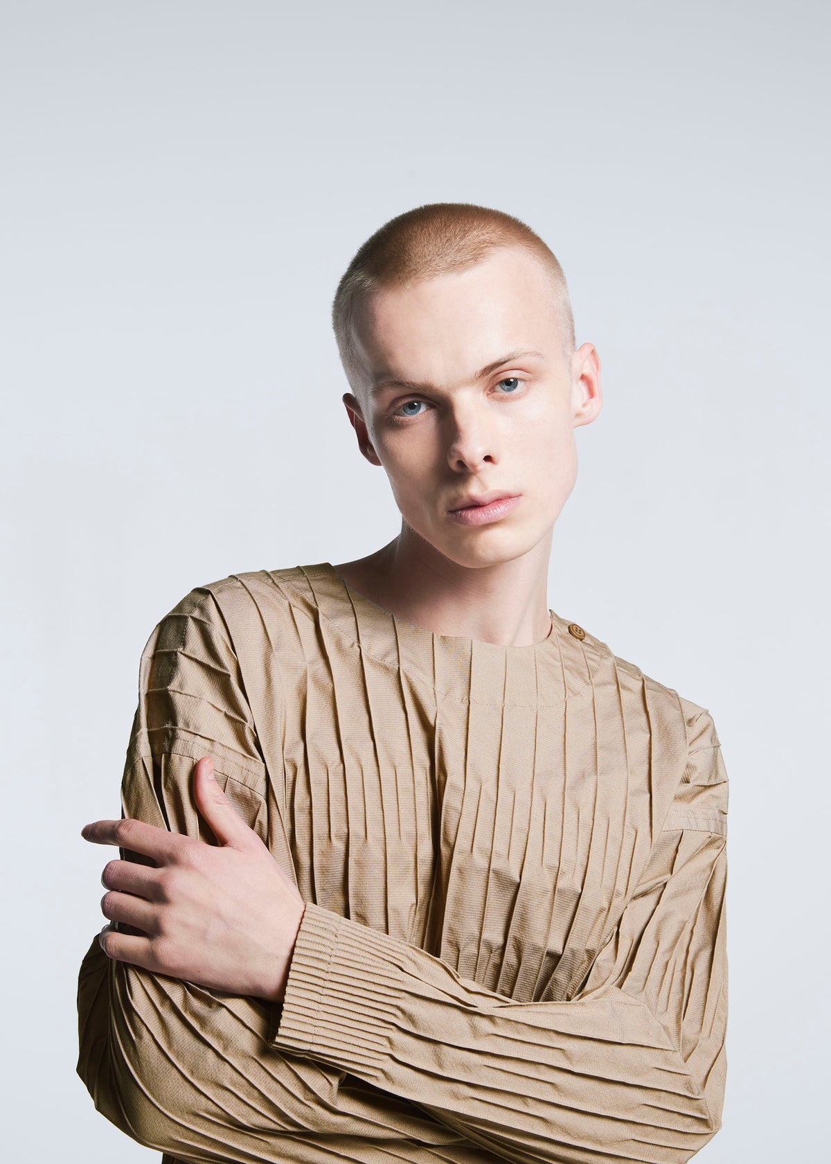A person with a shaved head models the tan TYPE-W 007 top by A-POC ABLE ISSEY MIYAKE, featuring subtle stripes, standing against a light gray background, looking at the camera with a neutral expression and one arm crossed. view 6 of 6