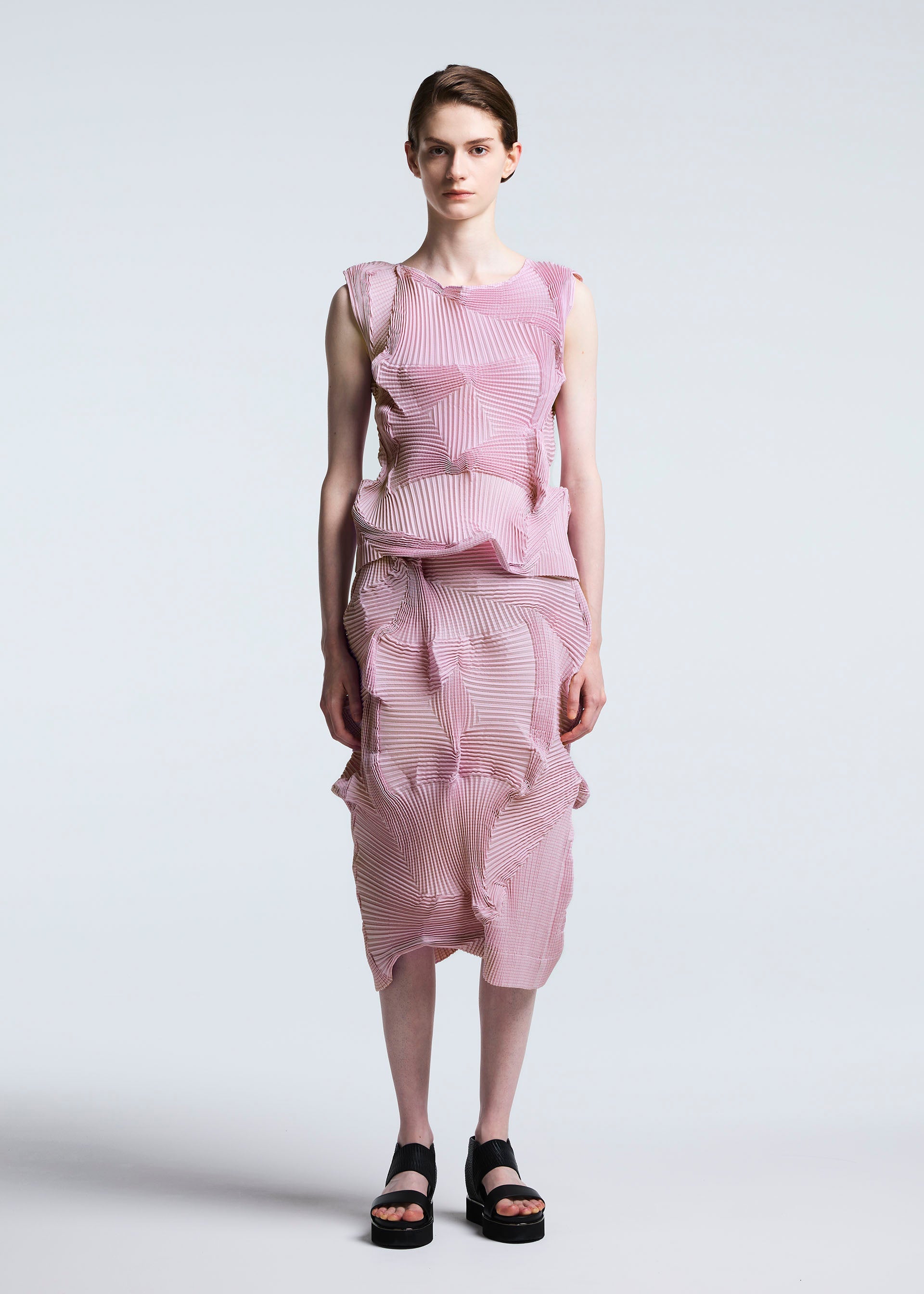 Zoomed view A person with short hair stands against a light background, wearing the sleeveless pink and white striped Steam Stretch set with textured wavy details, paired with TYPE-O 003 BK black platform sandals by A-POC ABLE ISSEY MIYAKE.