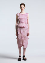 A person stands against a plain background, wearing TYPE-O 003 BK by A-POC ABLE ISSEY MIYAKE—a sleeveless pink and white striped steam stretch top with matching pleated midi skirt in recycled polyester, styled with black platform sandals and short brown hair.