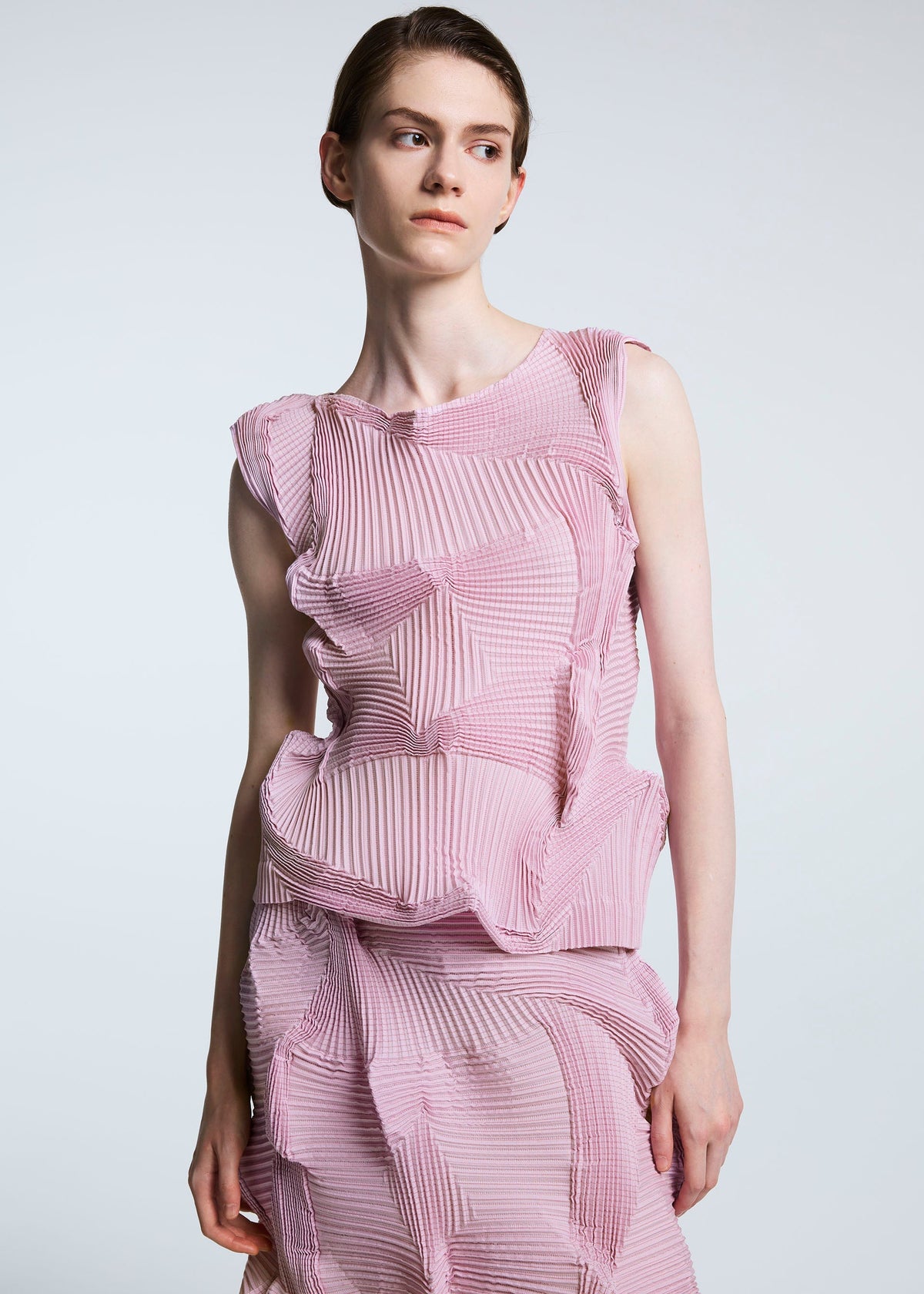 Wearing the TYPE-O 003 BK by A-POC ABLE ISSEY MIYAKE, a person in a sleeveless pink recycled polyester top and a geometric textured skirt stands against a plain light background, gazing slightly to the side. view 3 of 4
