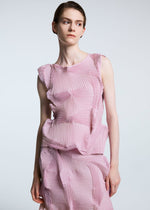 Wearing the TYPE-O 003 BK by A-POC ABLE ISSEY MIYAKE, a person in a sleeveless pink recycled polyester top and a geometric textured skirt stands against a plain light background, gazing slightly to the side.