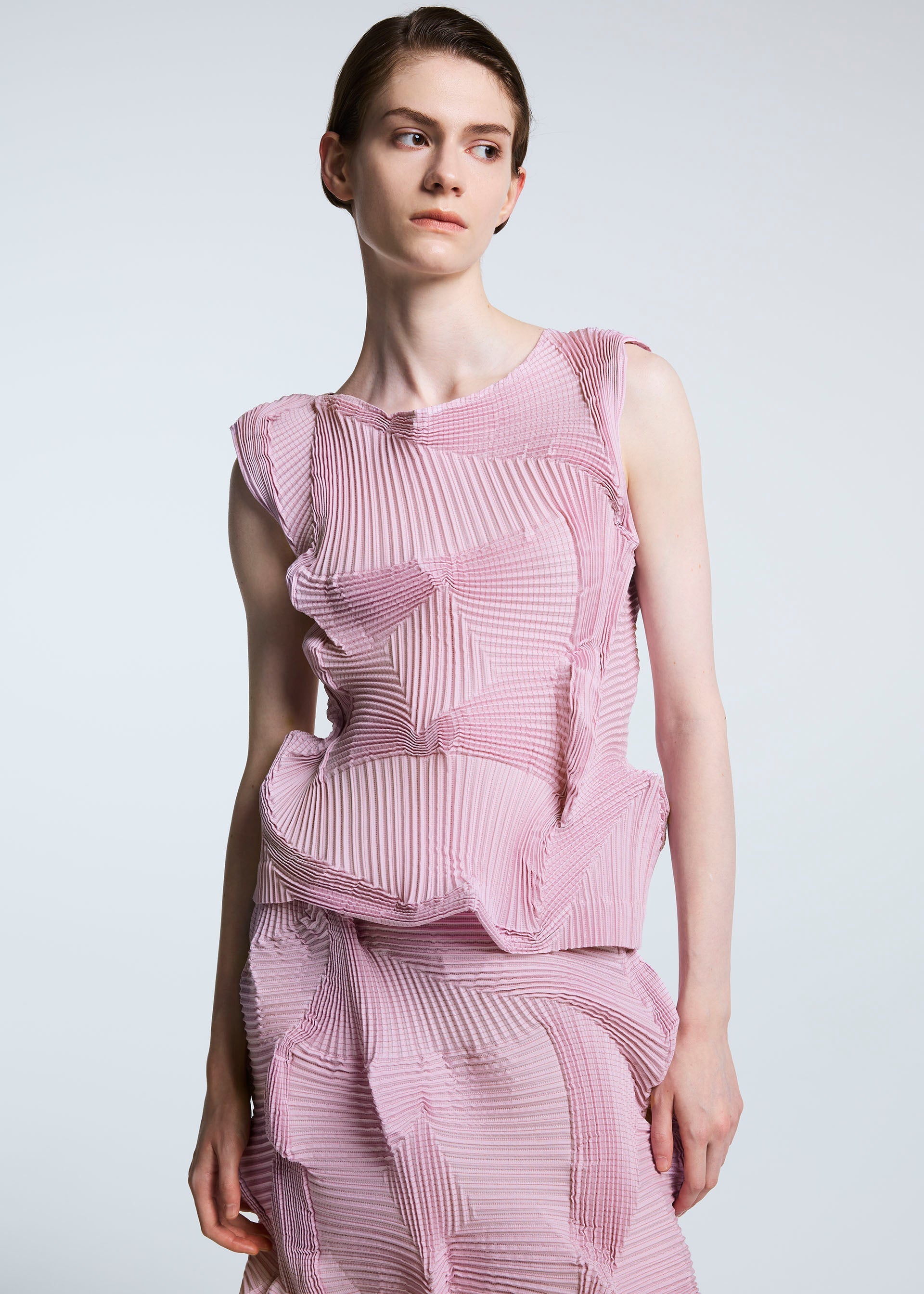 Zoomed view Wearing the TYPE-O 003 BK by A-POC ABLE ISSEY MIYAKE, a person in a sleeveless pink recycled polyester top and a geometric textured skirt stands against a plain light background, gazing slightly to the side.
