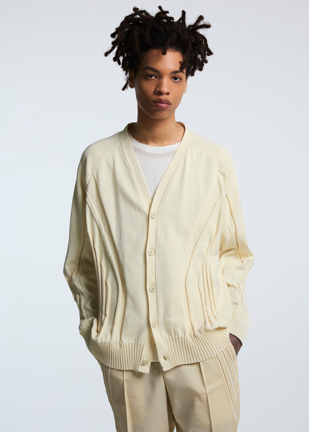 A person with short, curly hair wears the TYPE-K 003-1 cardigan by A-POC ABLE ISSEY MIYAKE over a white shirt and cream pants, standing against a plain light background with hands in pockets. view 5 of 7