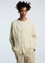 A person with short, curly hair wears the TYPE-K 003-1 cardigan by A-POC ABLE ISSEY MIYAKE over a white shirt and cream pants, standing against a plain light background with hands in pockets.