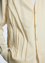 A close-up of the TYPE-K 003-1 cream knit cardigan by A-POC ABLE ISSEY MIYAKE, featuring a button front, ribbed cuffs, unique three-dimensional knitting, stitched side pleats, and a visible white shirt underneath.
