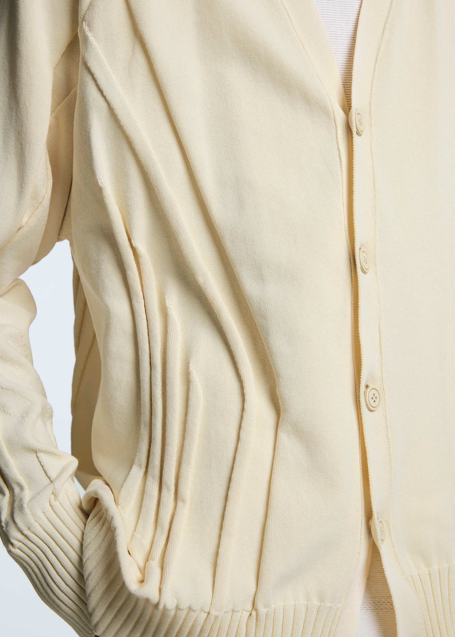 Zoomed view A close-up of the TYPE-K 003-1 cream knit cardigan by A-POC ABLE ISSEY MIYAKE, featuring a button front, ribbed cuffs, unique three-dimensional knitting, stitched side pleats, and a visible white shirt underneath.