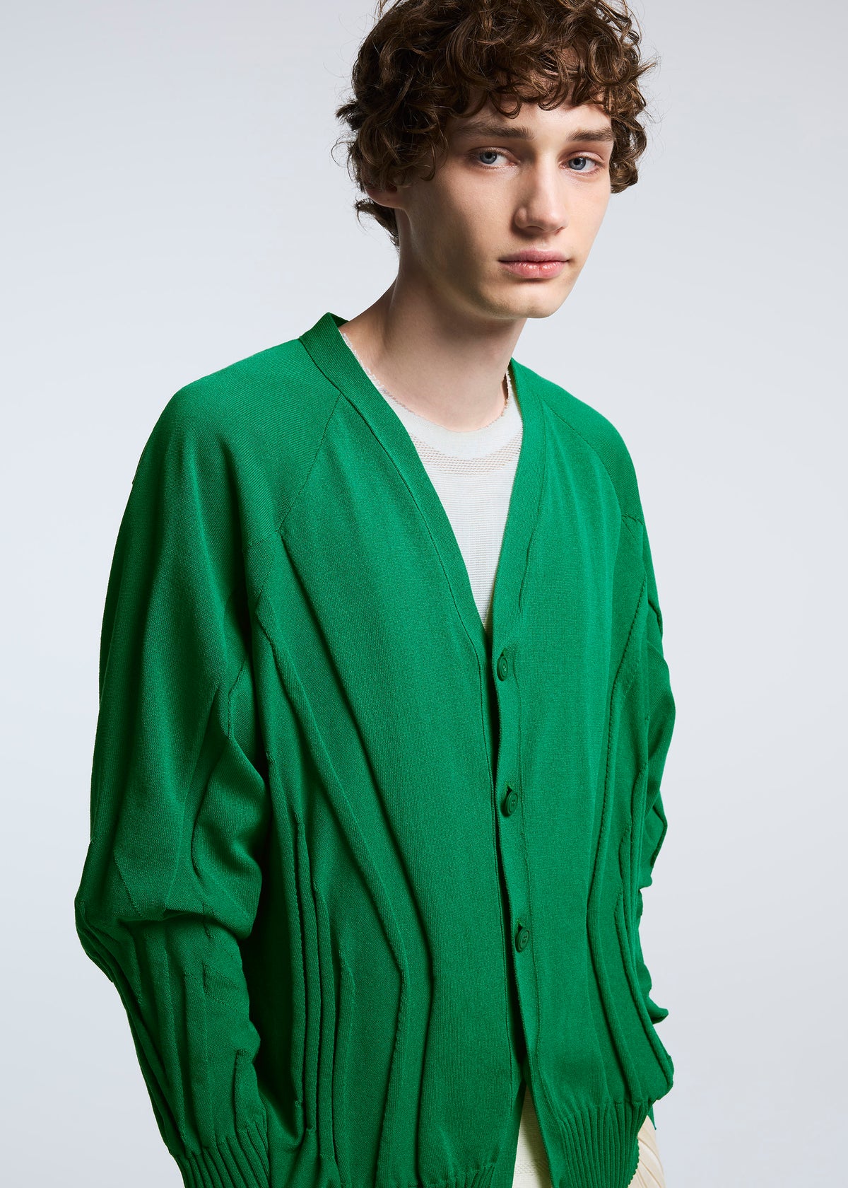 A young person with short curly brown hair wears the TYPE-K 003-1 bright green knit cardigan by A-POC ABLE ISSEY MIYAKE over a light shirt, standing against a plain light gray background. view 7 of 7