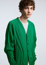 A young person with short curly brown hair wears the TYPE-K 003-1 bright green knit cardigan by A-POC ABLE ISSEY MIYAKE over a light shirt, standing against a plain light gray background.