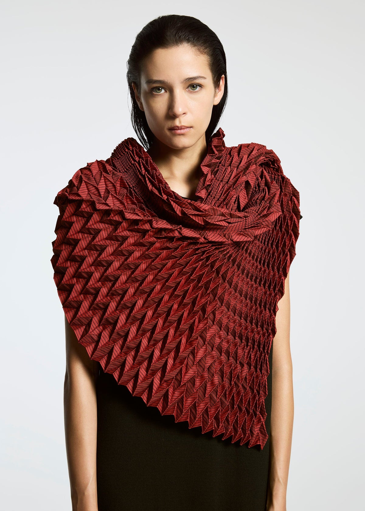 A person with straight, dark hair wears the TYPE-O STOLE by A-POC ABLE ISSEY MIYAKE in deep red, its sculptural three-dimensional pattern wrapping around their shoulders against a plain, light background that highlights the intricate fabric design. view 5 of 17