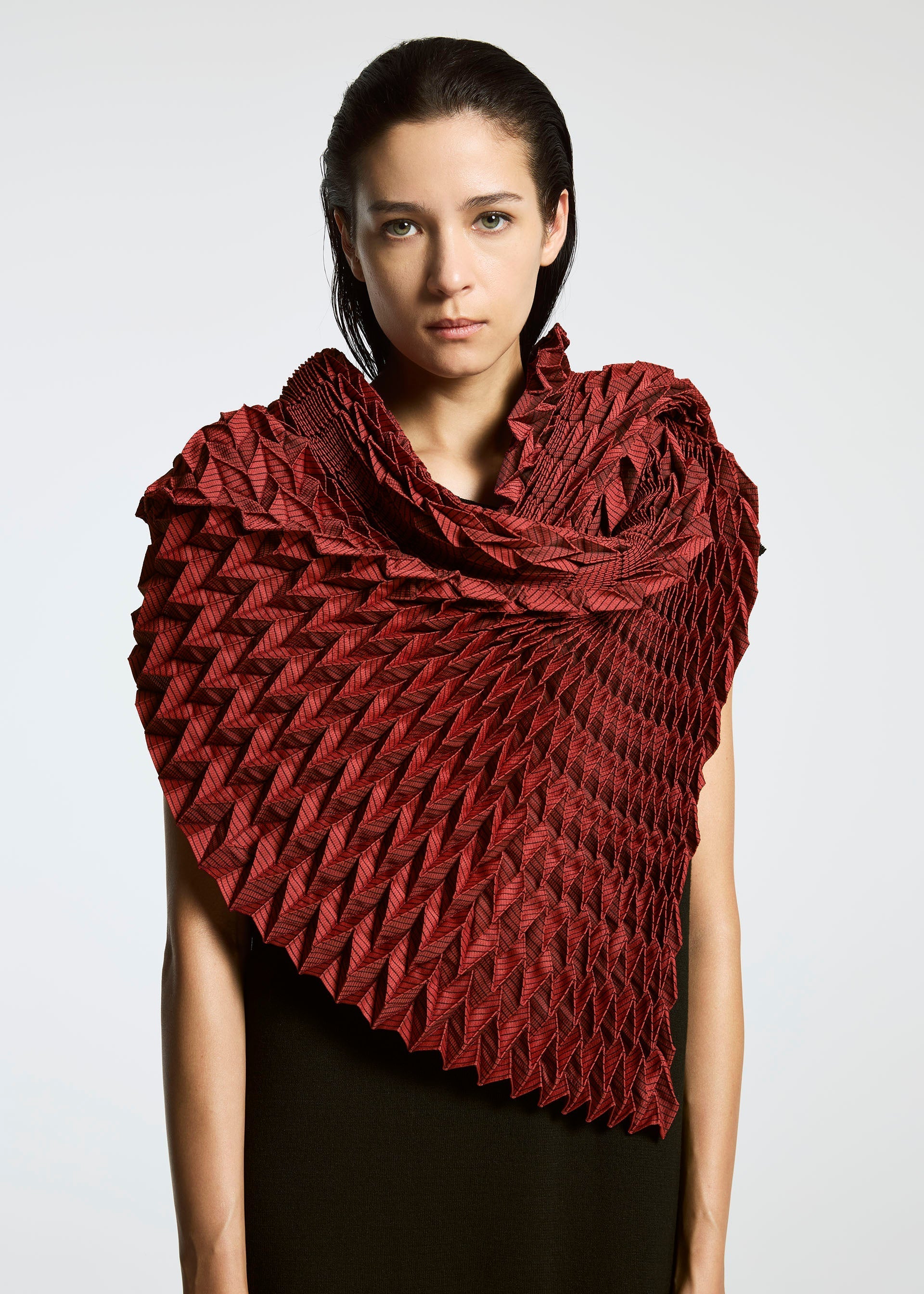 Zoomed view A person with straight, dark hair wears the TYPE-O STOLE by A-POC ABLE ISSEY MIYAKE in deep red, its sculptural three-dimensional pattern wrapping around their shoulders against a plain, light background that highlights the intricate fabric design.
