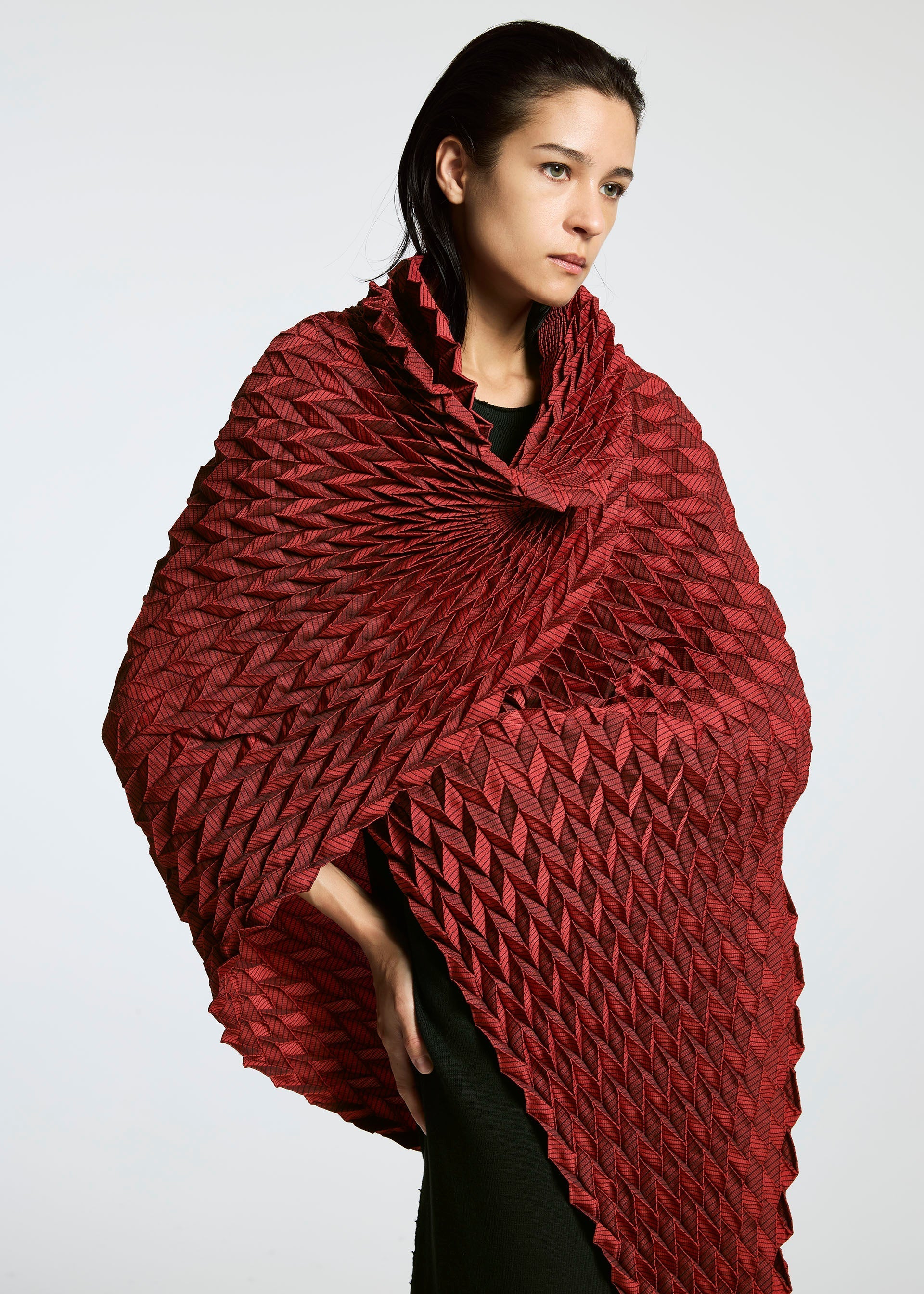 Zoomed view A woman stands against a plain white background, draped in the deep red TYPE-O STOLE by A-POC ABLE ISSEY MIYAKE, featuring a textured steam-stretch finish and a three-dimensional geometric pattern around her shoulders.