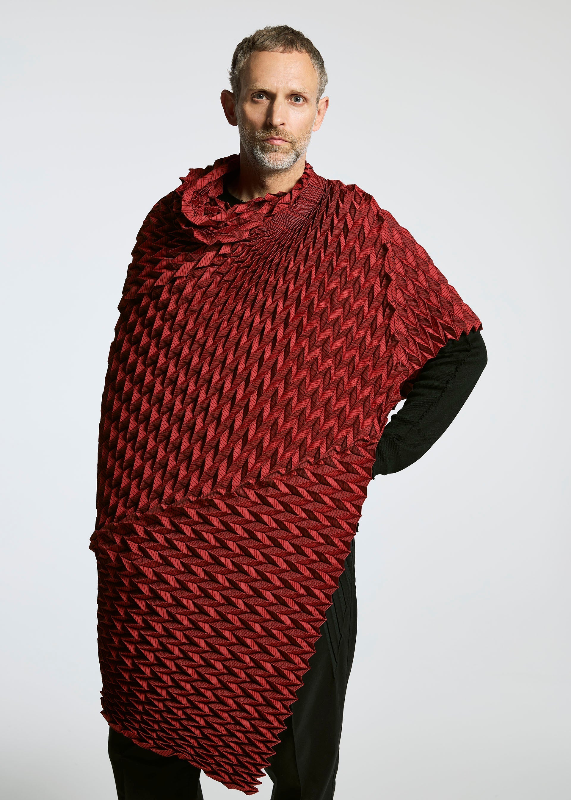 Zoomed view A person with short hair and a beard stands against a plain background, wearing the deep red TYPE-O STOLE by A-POC ABLE ISSEY MIYAKE—a sculptural piece with a 3D zigzag pattern—over a black outfit, accentuated by its voluminous design.