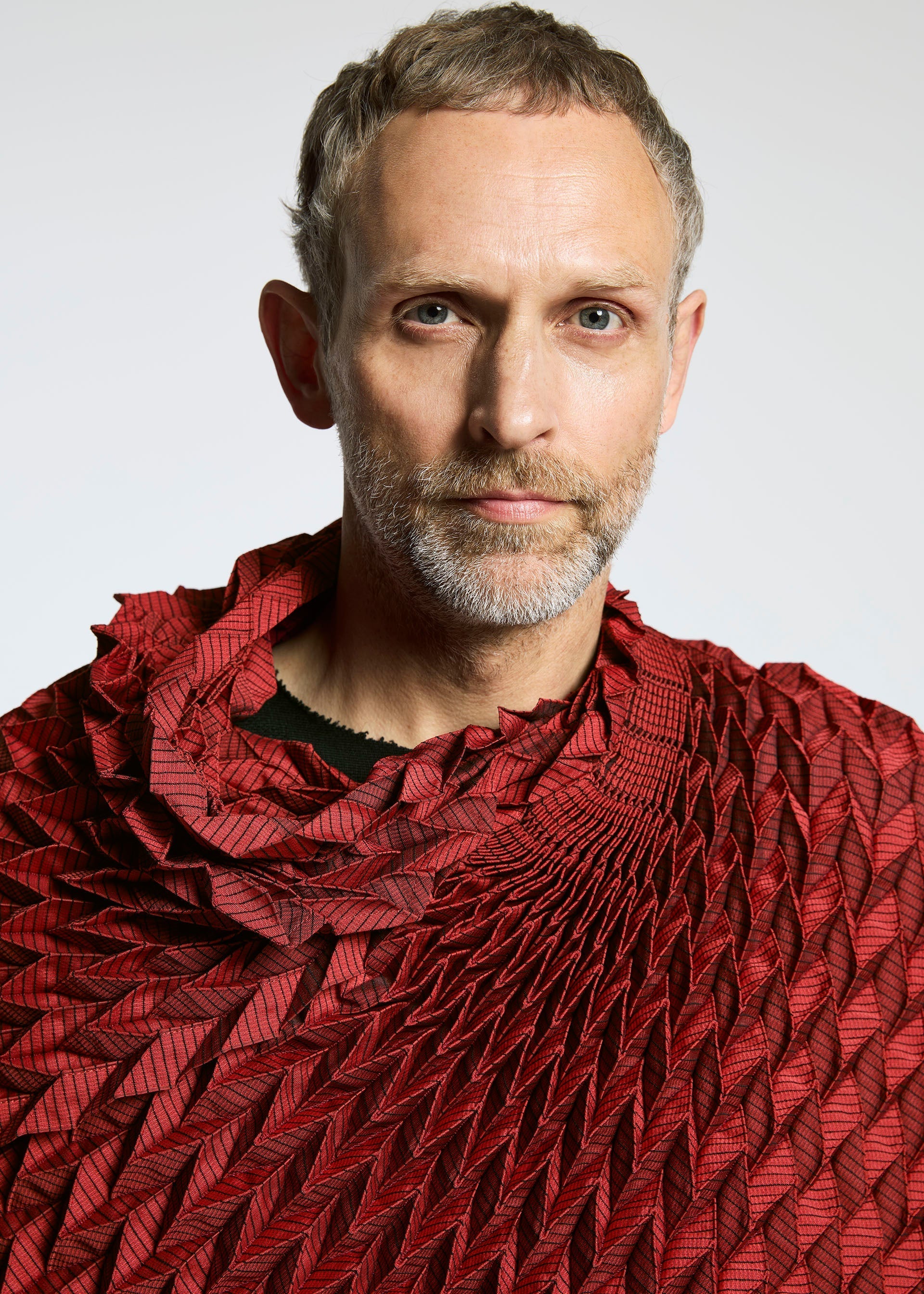 Zoomed view A man with short light hair and a beard wears the TYPE-O STOLE by A-POC ABLE ISSEY MIYAKE, a sculptural red garment with a three-dimensional pattern. He looks directly at the camera against a plain light background.