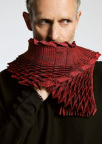 A man with light hair and beard holds the TYPE-O STOLE by A-POC ABLE ISSEY MIYAKE—a voluminous scarf with a geometric 3D pattern—over his mouth, dressed in black and looking to the side against a plain background.