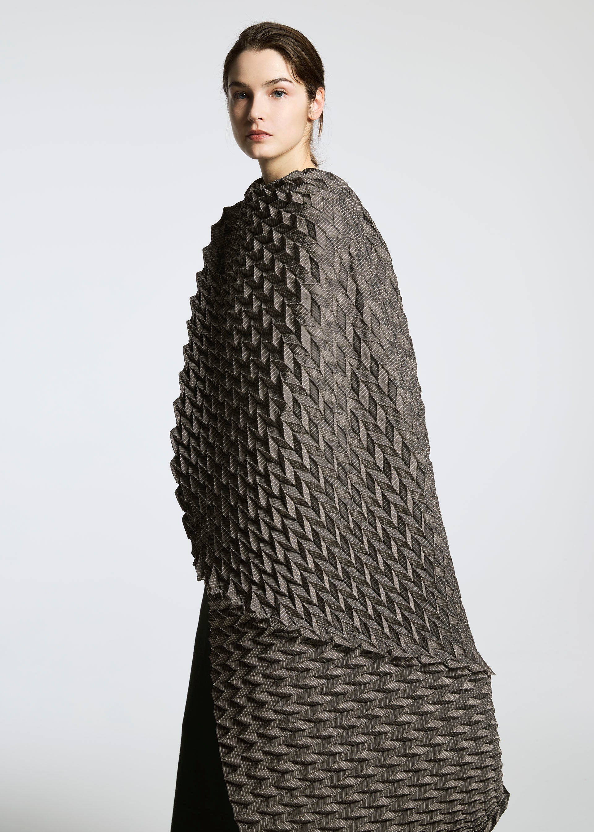 Zoomed view A woman models the TYPE-O STOLE by A-POC ABLE ISSEY MIYAKE—a textured, geometric-patterned gray and black cape with a voluminous scarf—looking at the camera against a plain light gray background.