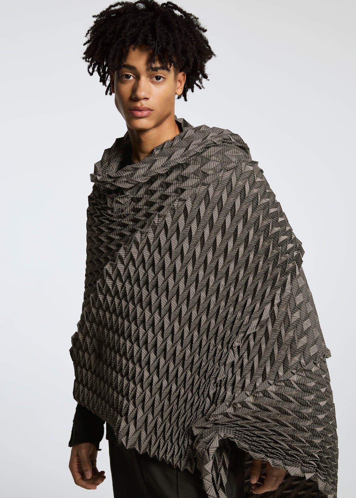A person with short, curly hair wears the gray TYPE-O STOLE by A-POC ABLE ISSEY MIYAKE, featuring a three-dimensional pattern, standing before a plain light background and looking calmly at the camera. view 12 of 17