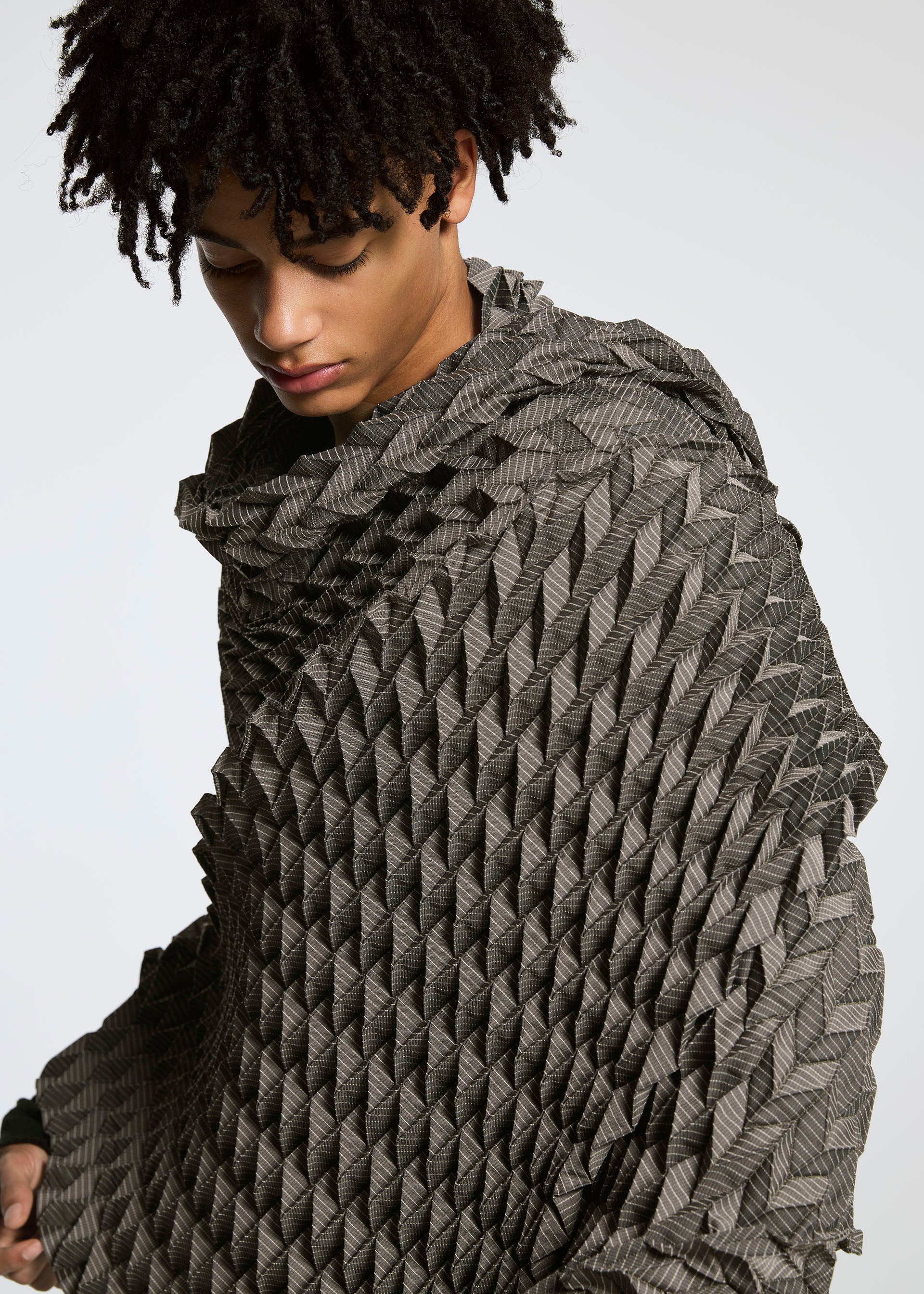 Zoomed view A person with short, curly hair wears the TYPE-O STOLE by A-POC ABLE ISSEY MIYAKE—a textured, geometric-patterned gray piece styled as a voluminous scarf—while looking downward against a plain light background.