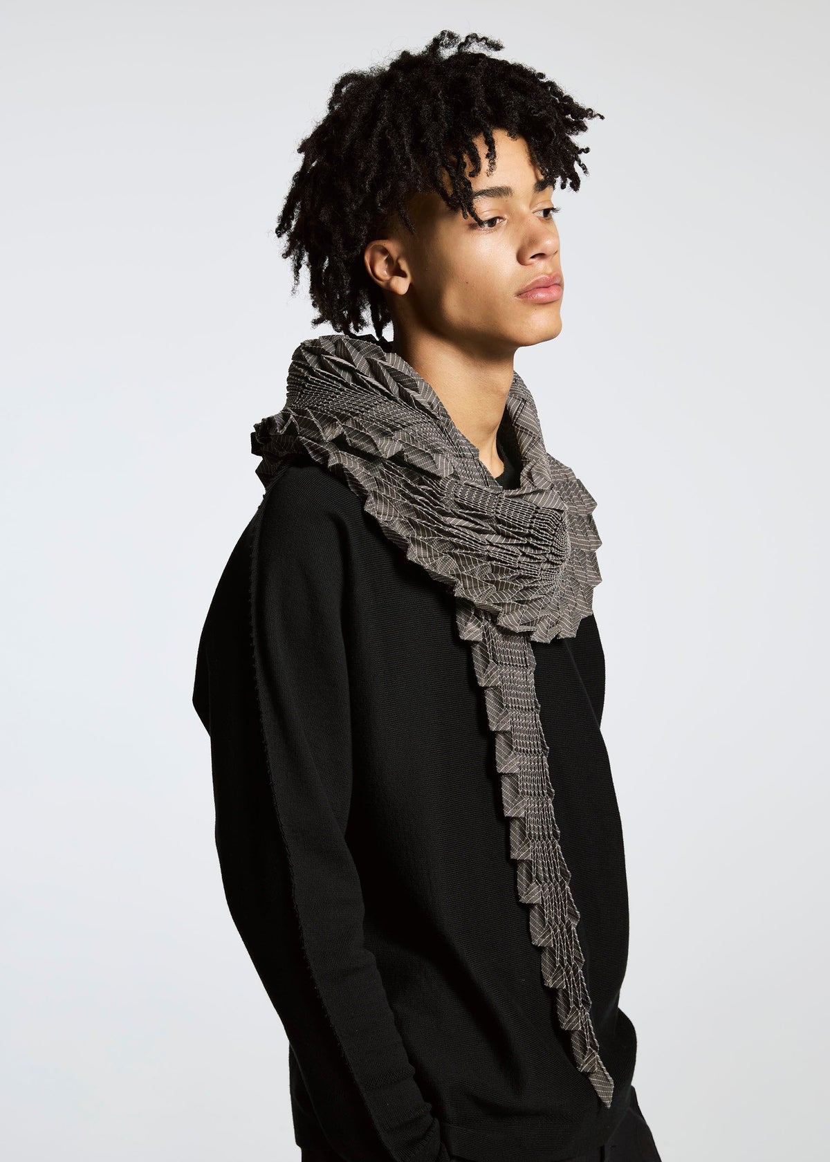 A person with short curly hair models the TYPE-O STOLE by A-POC ABLE ISSEY MIYAKE, featuring a voluminous 3D-patterned scarf and a black sweater, standing against a plain light background with a neutral expression. view 14 of 17