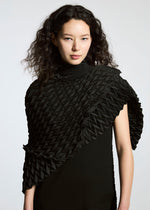 A person with wavy dark hair wears the TYPE-O STOLE by A-POC ABLE ISSEY MIYAKE, a textured black accessory with intricate, origami-like folds draped over one shoulder, resembling a voluminous scarf, set against a plain light background.