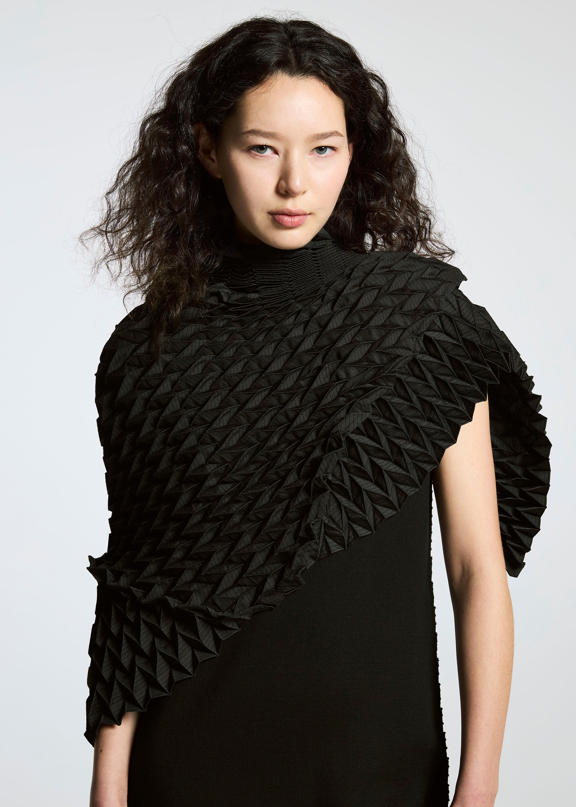 Zoomed view A person with wavy dark hair wears the TYPE-O STOLE by A-POC ABLE ISSEY MIYAKE, a textured black accessory with intricate, origami-like folds draped over one shoulder, resembling a voluminous scarf, set against a plain light background.