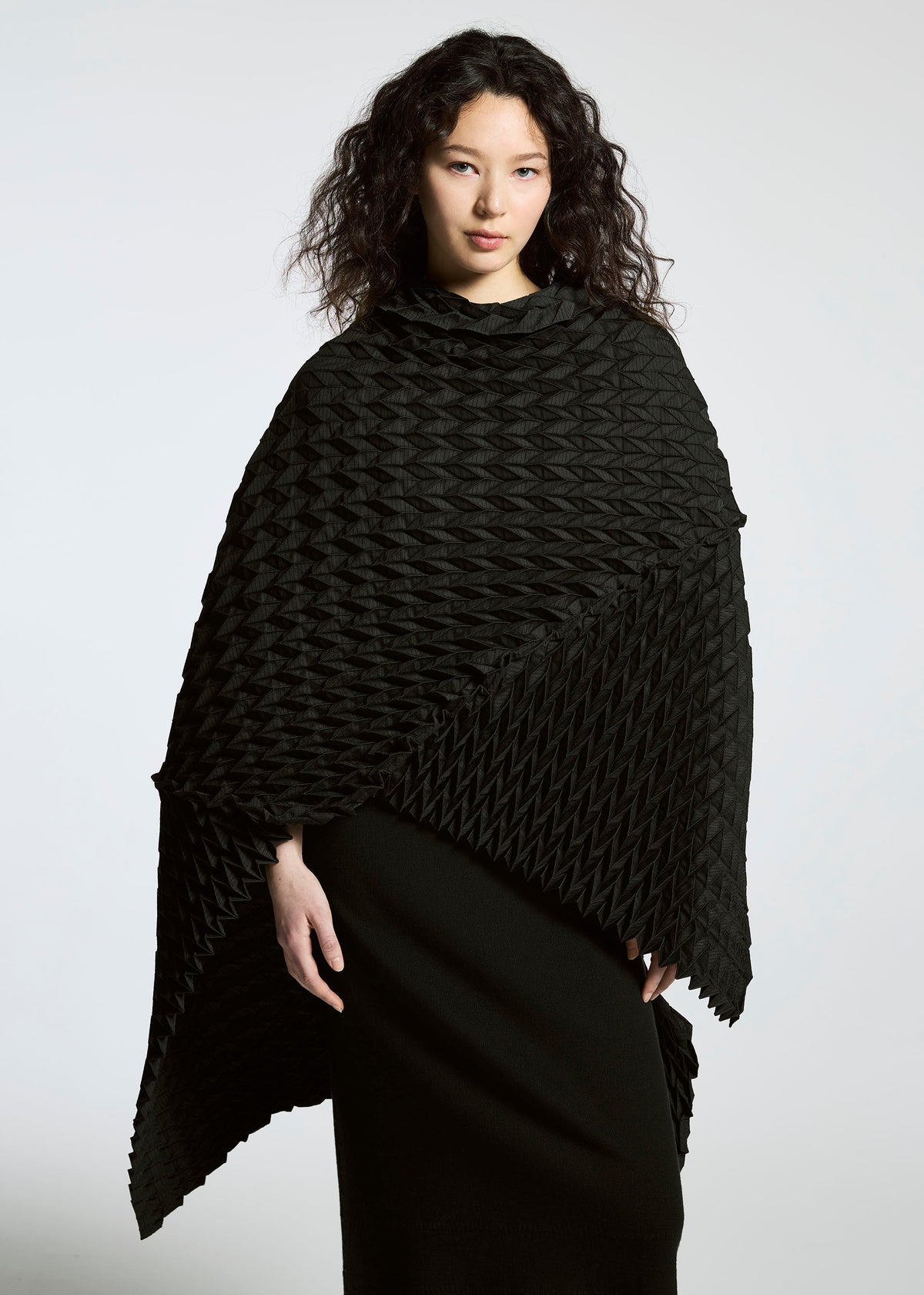 A woman with wavy dark hair wears the TYPE-O STOLE by A-POC ABLE ISSEY MIYAKE, a black textured poncho with a three-dimensional pattern, over a long black skirt against a plain light background. view 17 of 17