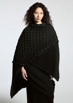 A woman with wavy dark hair wears the TYPE-O STOLE by A-POC ABLE ISSEY MIYAKE, a black textured poncho with a three-dimensional pattern, over a long black skirt against a plain light background.