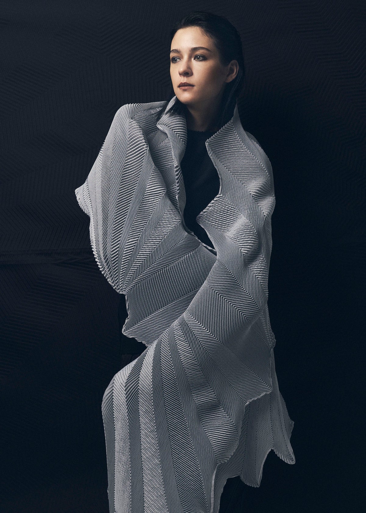 A woman stands against a dark background, wrapped in the TYPE-XIV Eugene Studio project 002 shawl by A-POC ABLE ISSEY MIYAKE. She gazes upward with a calm expression, embodying the spirit of Light and Shadow Inside Me. view 6 of 11
