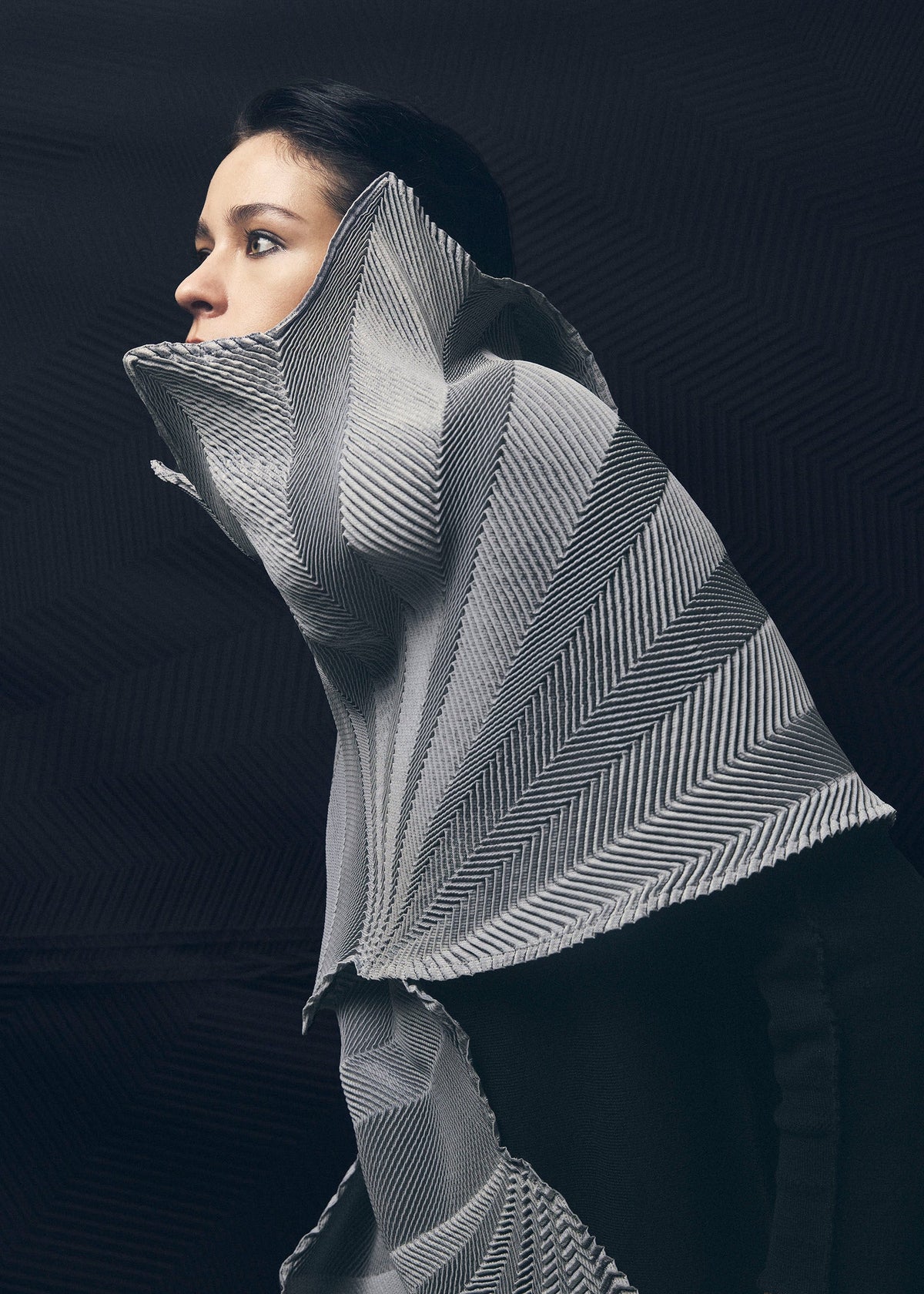 A woman with dark hair wears A-POC ABLE ISSEY MIYAKE's TYPE-XIV Eugene Studio project 002, a sculptural, high-collared garment in black-and-white chevron, set against a dark background, evoking the interplay of Light and Shadow Inside Me. view 7 of 11