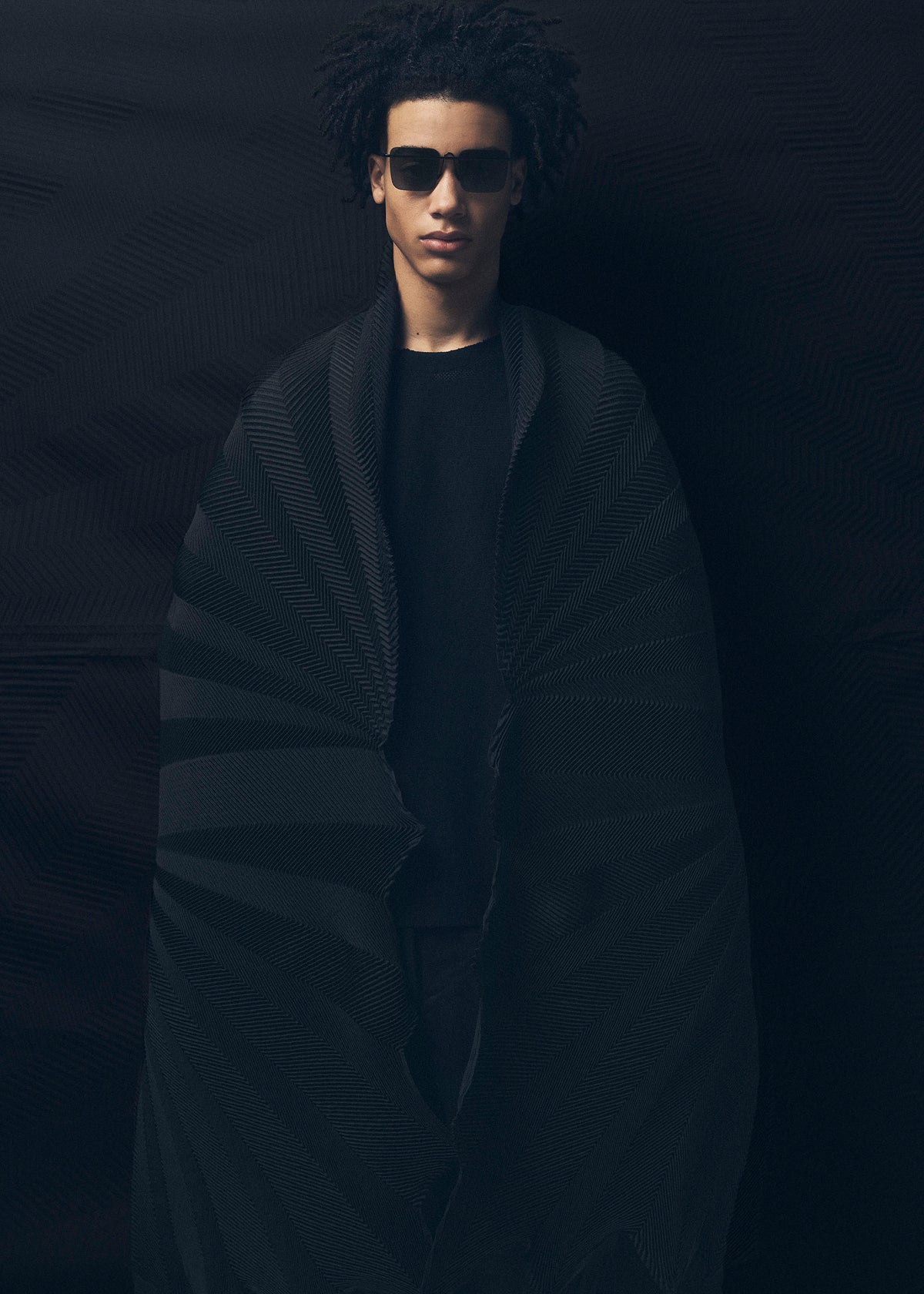 A person with short curly hair and dark sunglasses wears an oversized TYPE-XIV Eugene Studio project 002 cape by A-POC ABLE ISSEY MIYAKE, standing against a dark background. Dramatic lighting evokes Eugene Studio’s Light and Shadow Inside Me. view 8 of 11