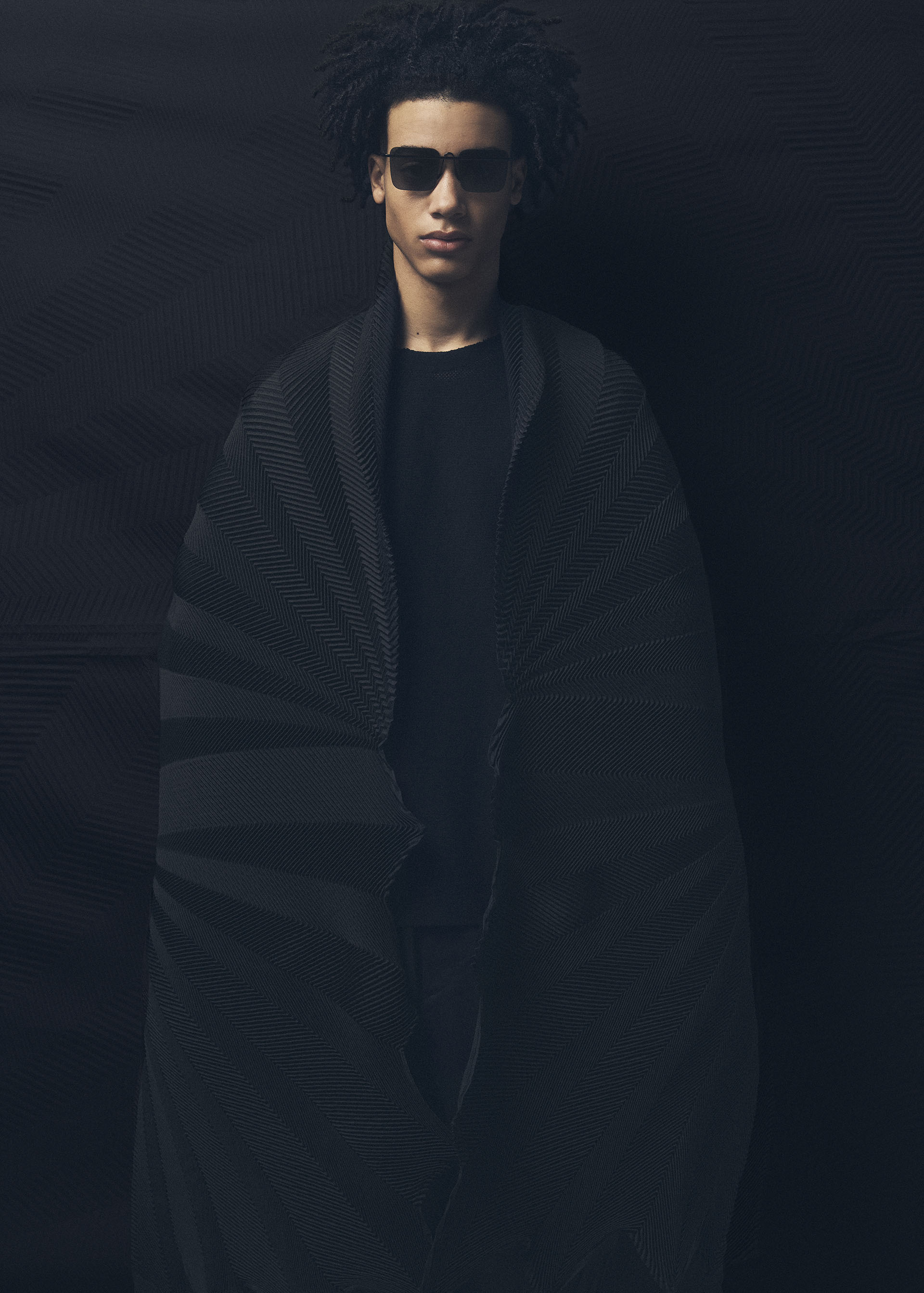 Zoomed view A person with short curly hair and dark sunglasses wears an oversized TYPE-XIV Eugene Studio project 002 cape by A-POC ABLE ISSEY MIYAKE, standing against a dark background. Dramatic lighting evokes Eugene Studio’s Light and Shadow Inside Me.