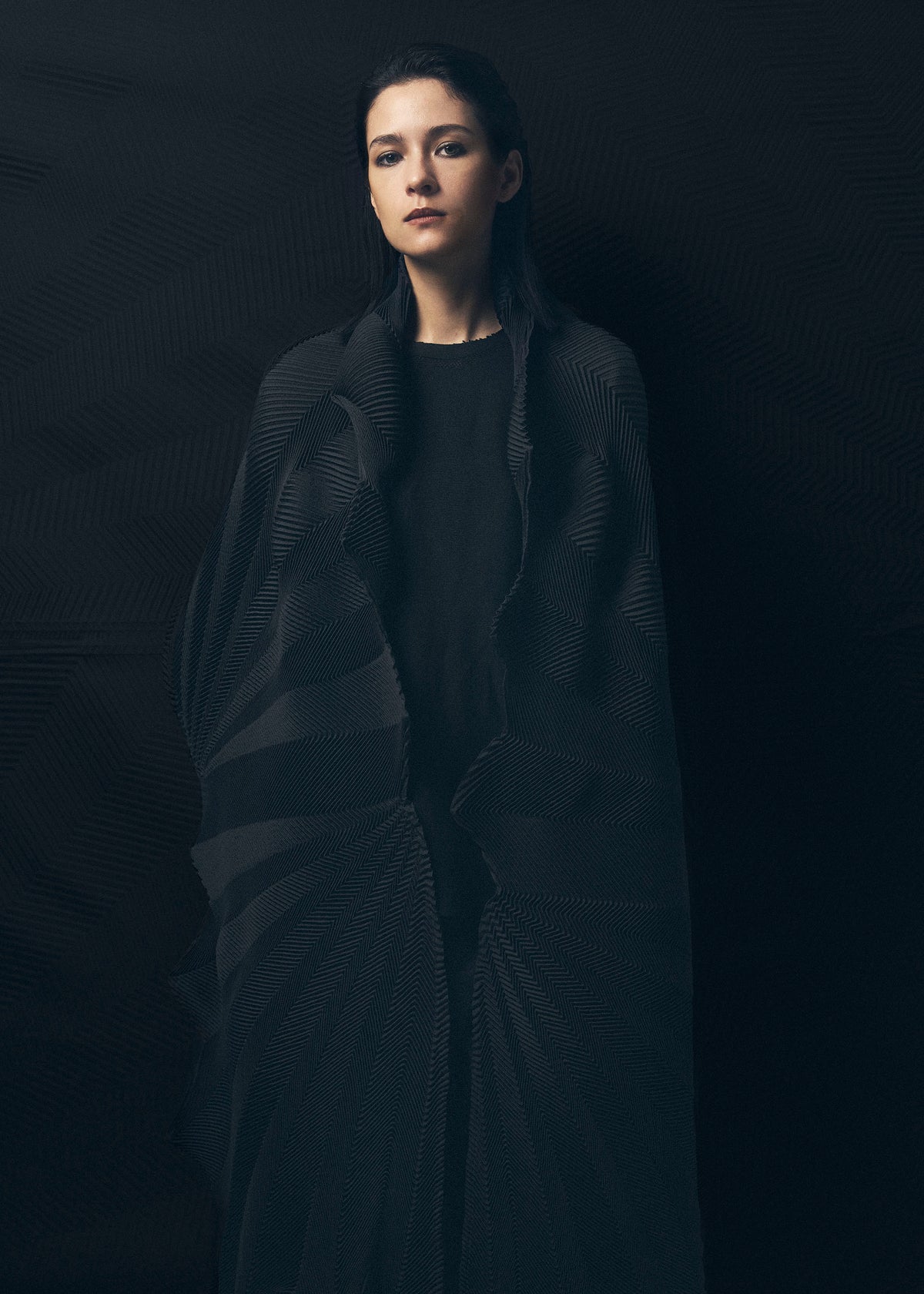A young woman with straight dark hair stands against a dark background, draped in the TYPE-XIV Eugene Studio project 002 by A-POC ABLE ISSEY MIYAKE, gazing forward with a calm, neutral expression. view 9 of 11