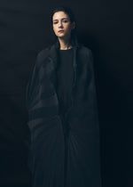 A young woman with straight dark hair stands against a dark background, draped in the TYPE-XIV Eugene Studio project 002 by A-POC ABLE ISSEY MIYAKE, gazing forward with a calm, neutral expression.