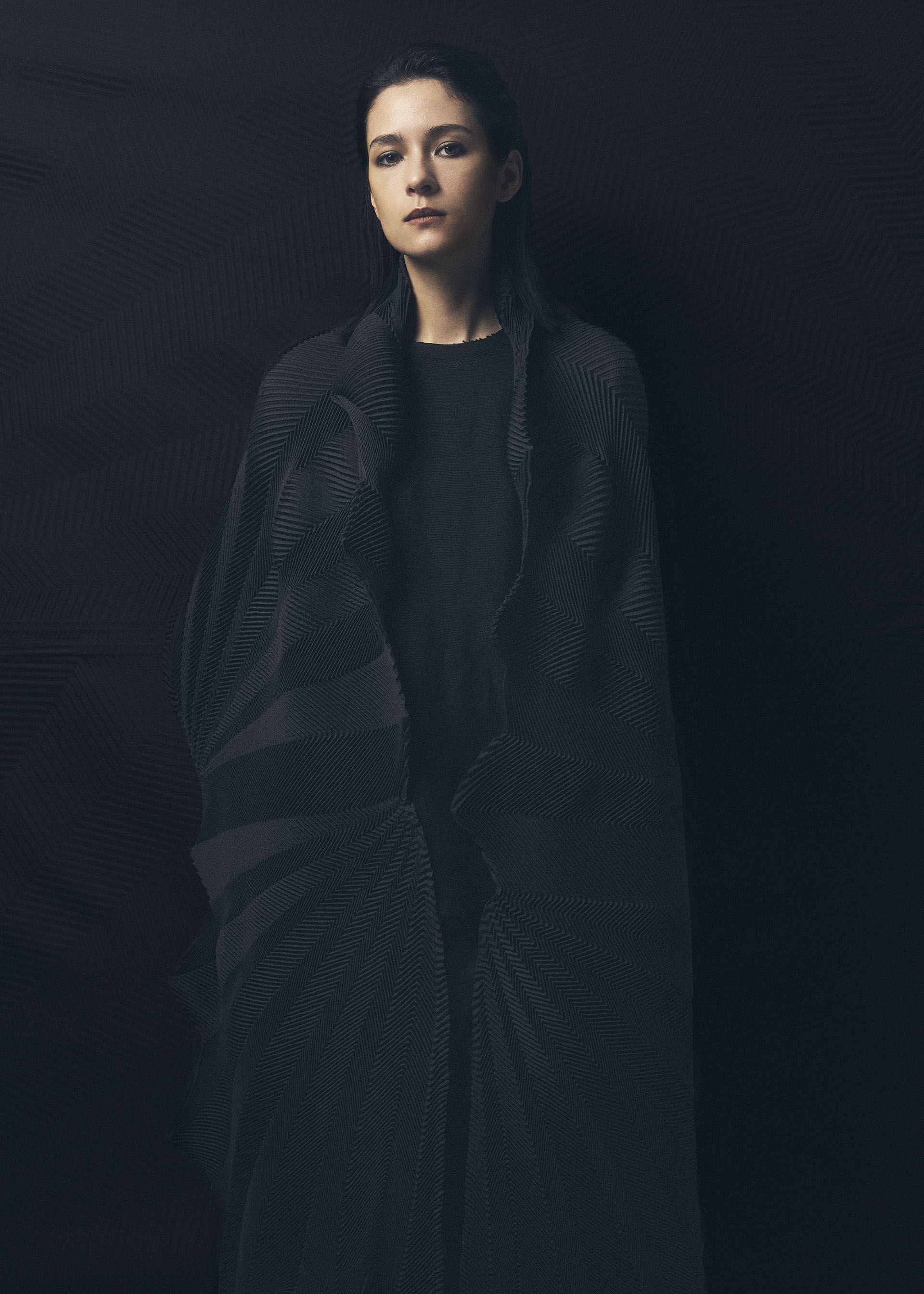 Zoomed view A young woman with straight dark hair stands against a dark background, draped in the TYPE-XIV Eugene Studio project 002 by A-POC ABLE ISSEY MIYAKE, gazing forward with a calm, neutral expression.