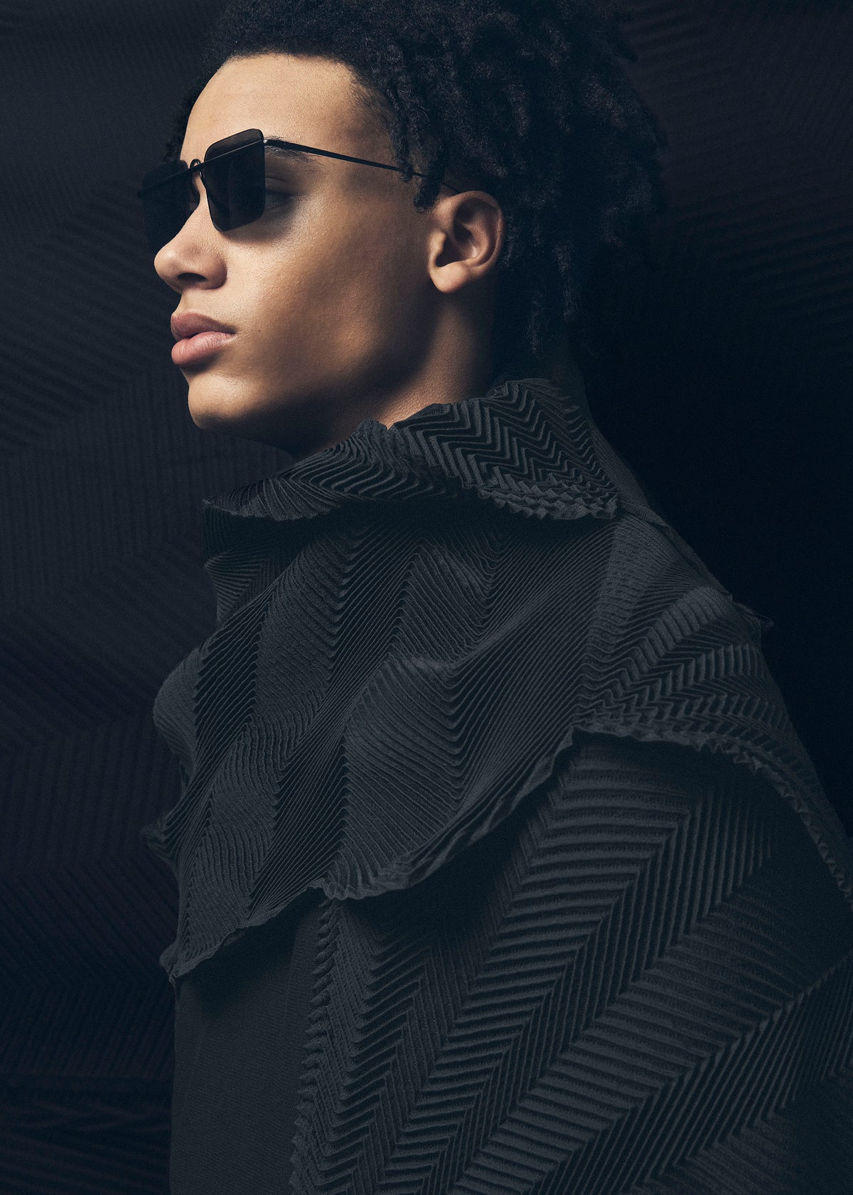 A person with short curly hair wears dark sunglasses and a textured, patterned black TYPE-XIV Eugene Studio project 002 by A-POC ABLE ISSEY MIYAKE, looking to the left against a dark, rippled background. view 10 of 11