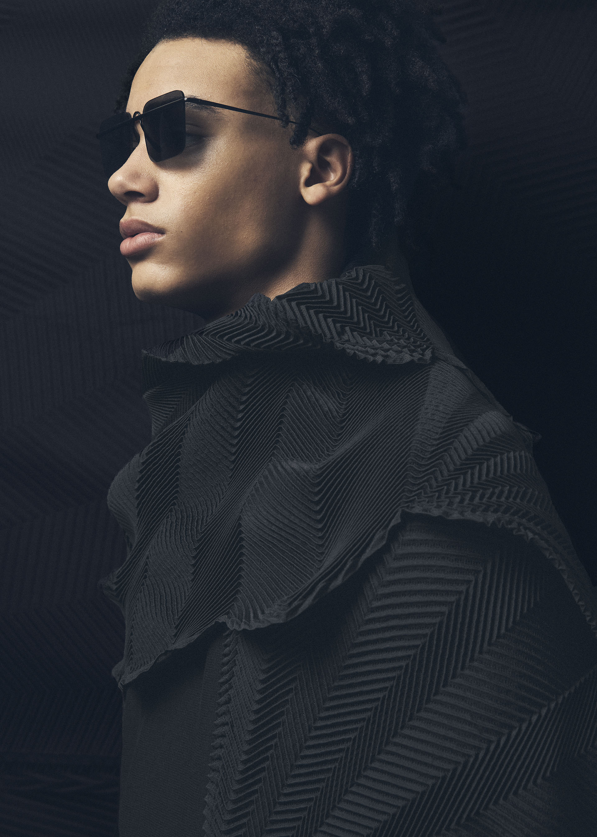 Zoomed view A person with short curly hair wears dark sunglasses and a textured, patterned black TYPE-XIV Eugene Studio project 002 by A-POC ABLE ISSEY MIYAKE, looking to the left against a dark, rippled background.