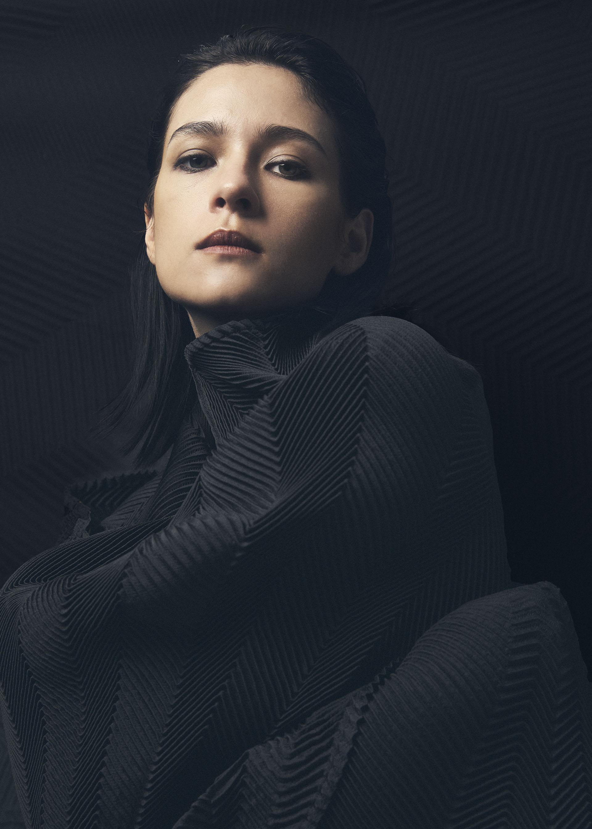 Zoomed view A person with dark hair and a serious expression wears the textured, pleated TYPE-XIV Eugene Studio project 002 by A-POC ABLE ISSEY MIYAKE against a dark background. Soft lighting highlights the theme “Light and Shadow Inside Me” and the garment’s details.