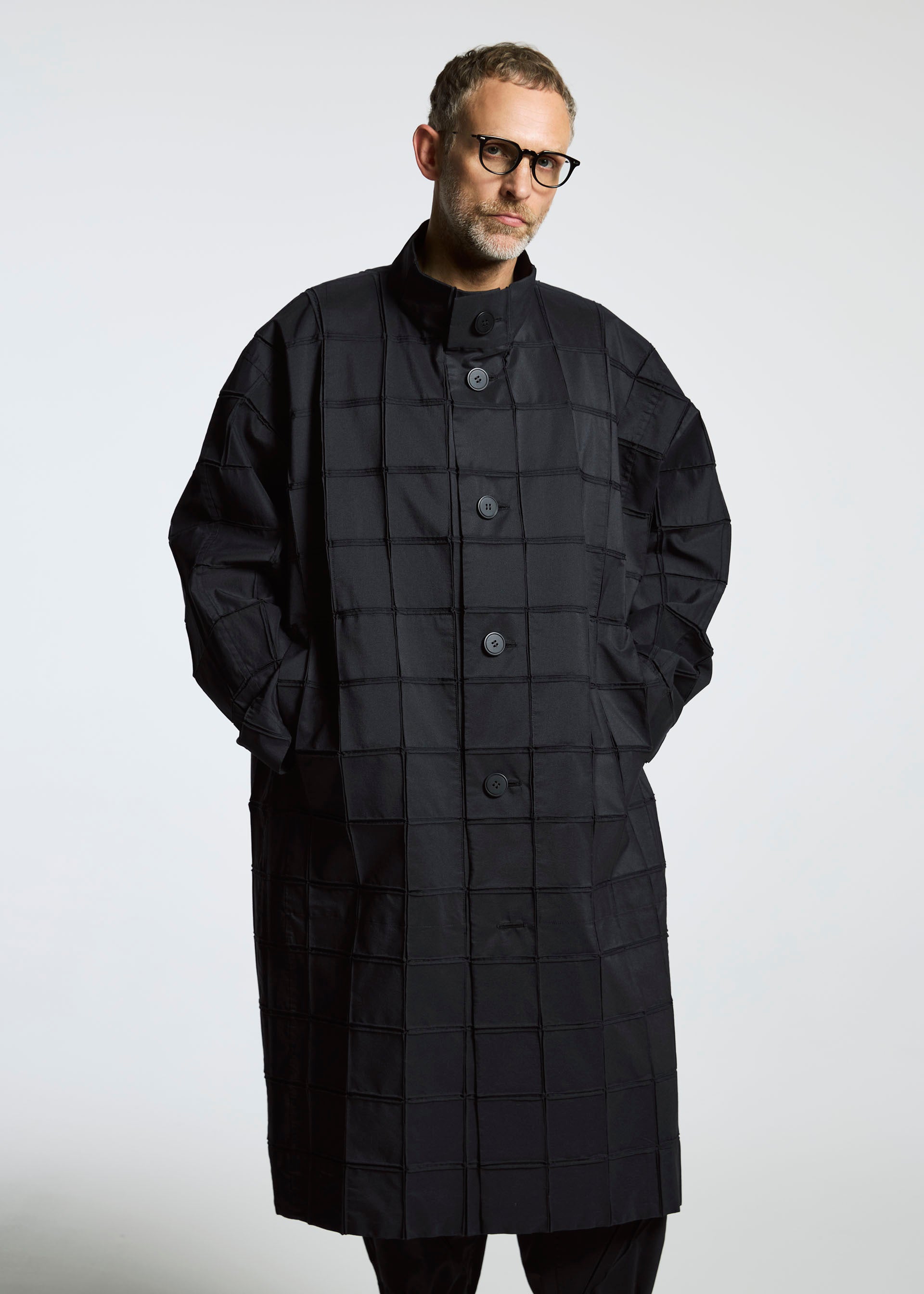 Zoomed view A man wearing glasses models the TYPE-O 002, a long, oversized grid-pattern Steam Stretch coat from A-POC ABLE ISSEY MIYAKE, standing with his hands in his pockets against a plain white background.