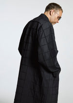 A man in glasses and a long, black grid coat from A-POC ABLE ISSEY MIYAKE’s TYPE-O 002 stands sideways, looking down against a plain light background.
