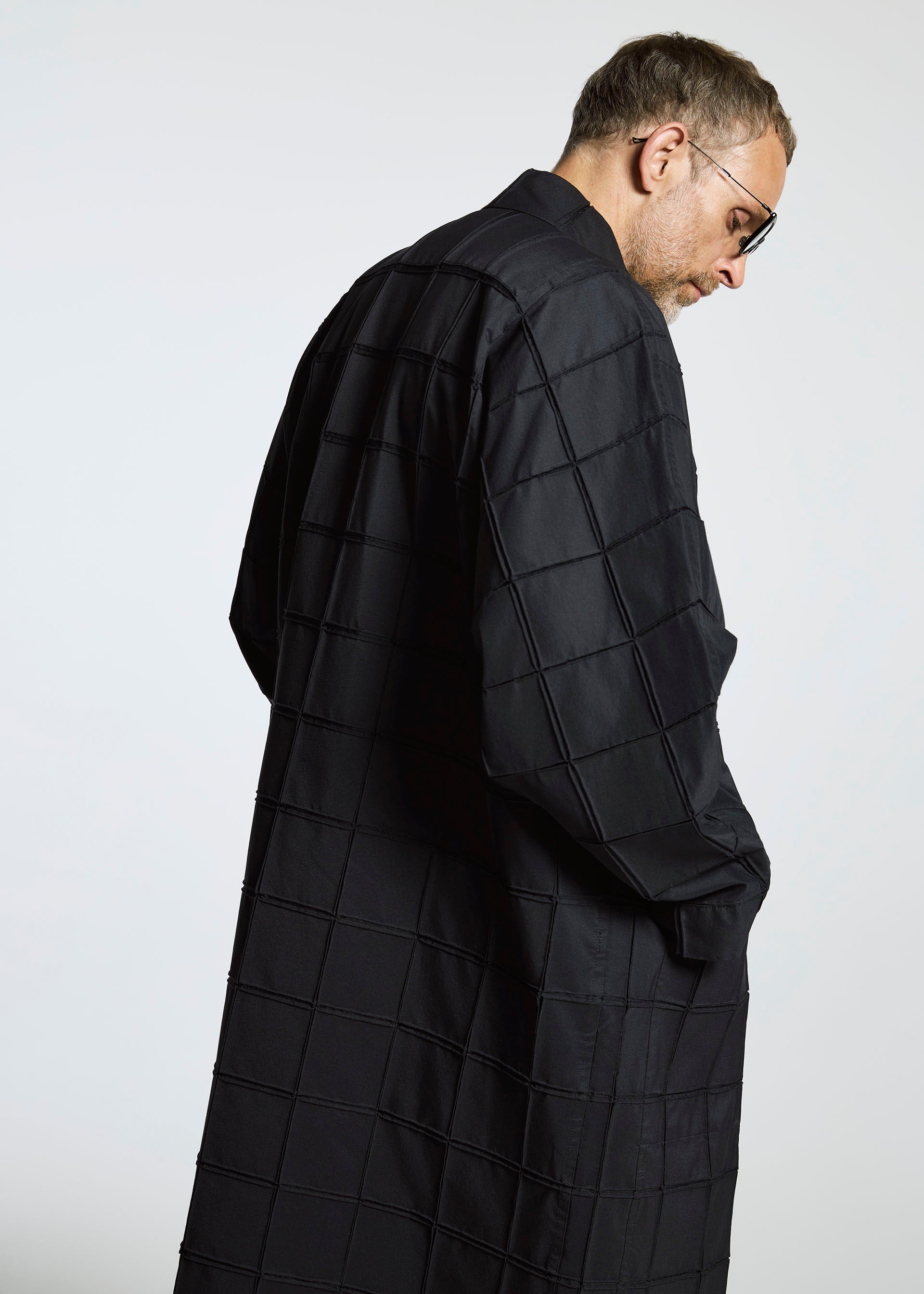 Zoomed view A man in glasses and a long, black grid coat from A-POC ABLE ISSEY MIYAKE’s TYPE-O 002 stands sideways, looking down against a plain light background.