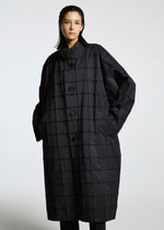 A person stands against a plain background wearing the TYPE-O 002 coat by A-POC ABLE ISSEY MIYAKE, featuring a loose fit, black grid pattern, oversized buttons, and hands in pockets while looking confidently at the camera.