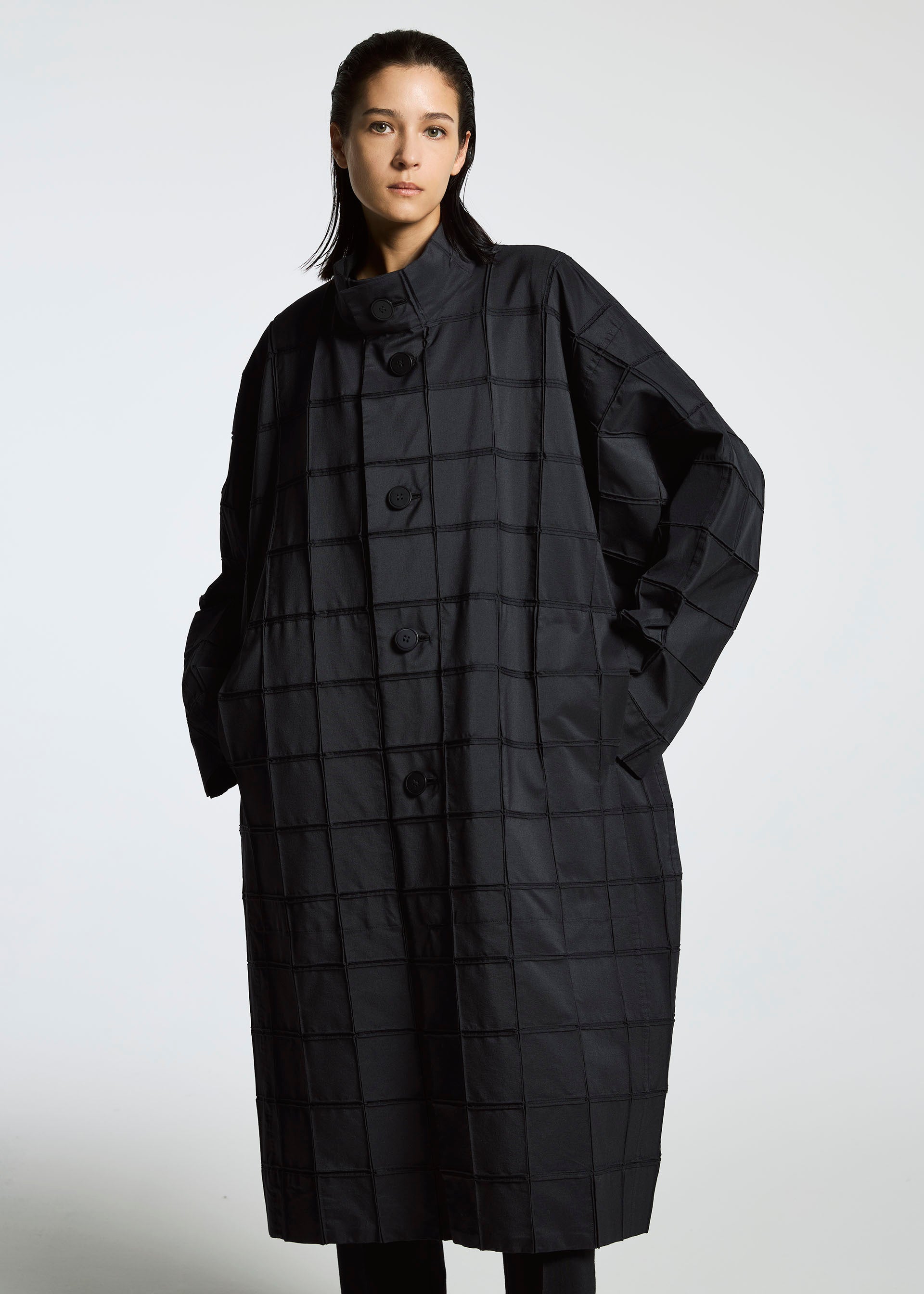 Zoomed view A person stands against a plain background wearing the TYPE-O 002 coat by A-POC ABLE ISSEY MIYAKE, featuring a loose fit, black grid pattern, oversized buttons, and hands in pockets while looking confidently at the camera.