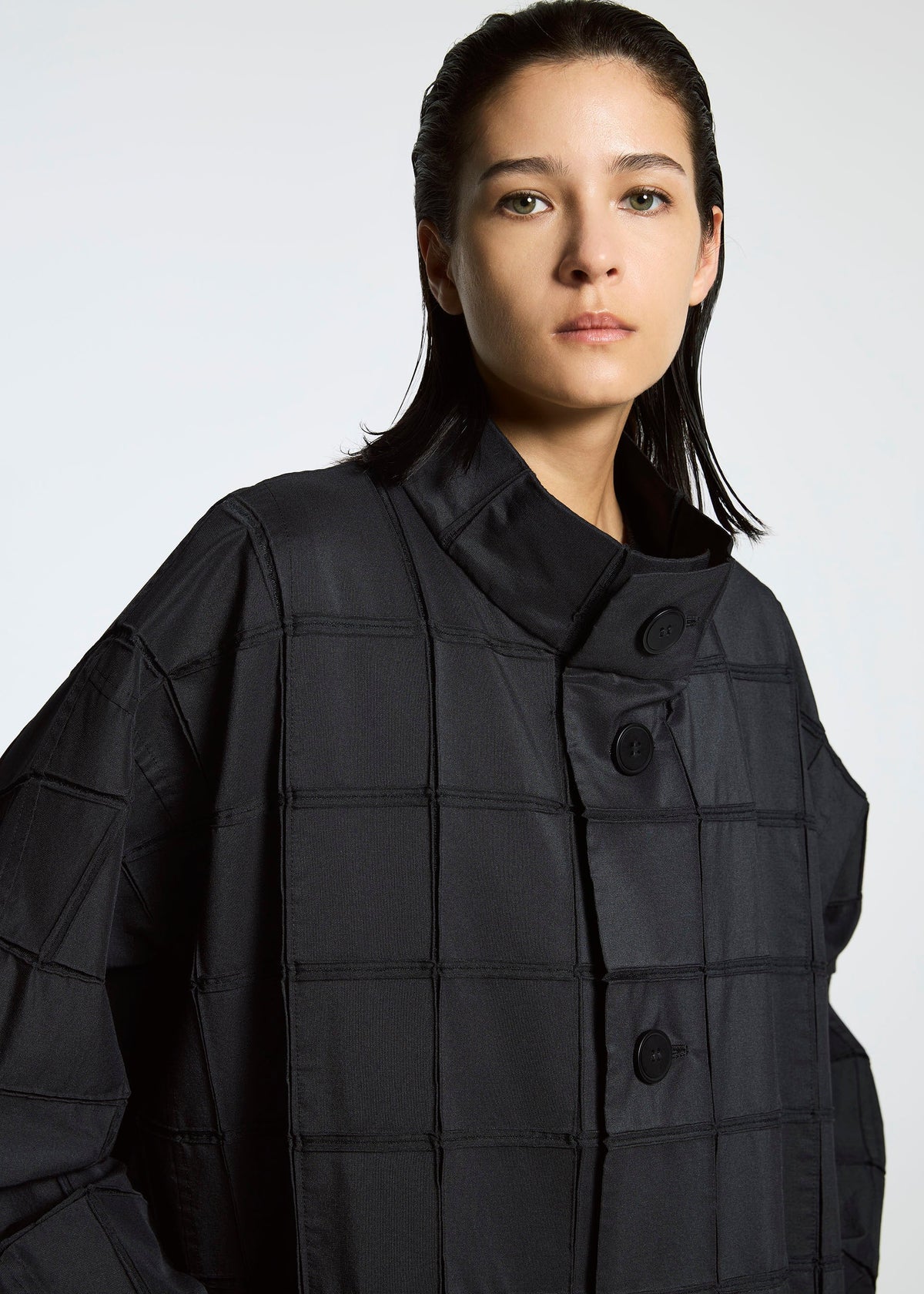 Wearing the TYPE-O 002 coat by A-POC ABLE ISSEY MIYAKE, a person with straight dark hair poses confidently for the camera, showcasing its grid pattern and large buttons against a plain background. view 8 of 10