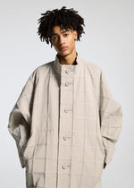 A person with short curly hair wears the TYPE-O 002 coat by A-POC ABLE ISSEY MIYAKE, featuring a light beige grid pattern and large buttons, hands in pockets, looking at the camera against a plain background.