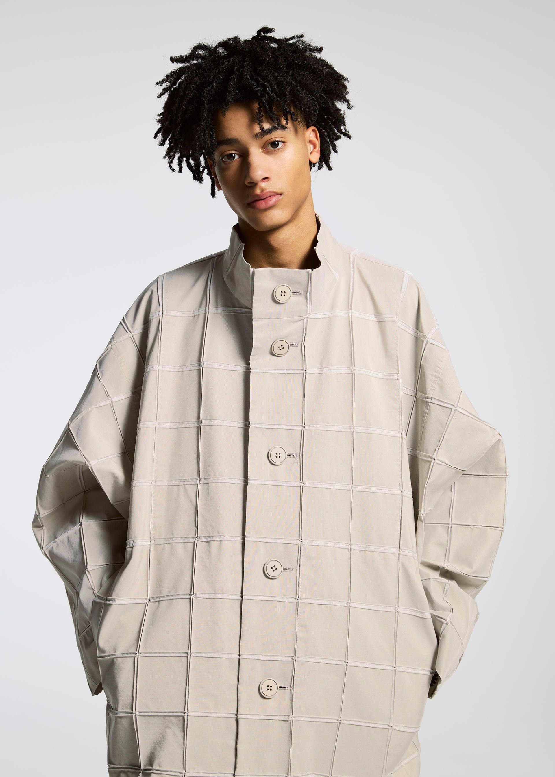 Zoomed view A person with short curly hair wears the TYPE-O 002 coat by A-POC ABLE ISSEY MIYAKE, featuring a light beige grid pattern and large buttons, hands in pockets, looking at the camera against a plain background.