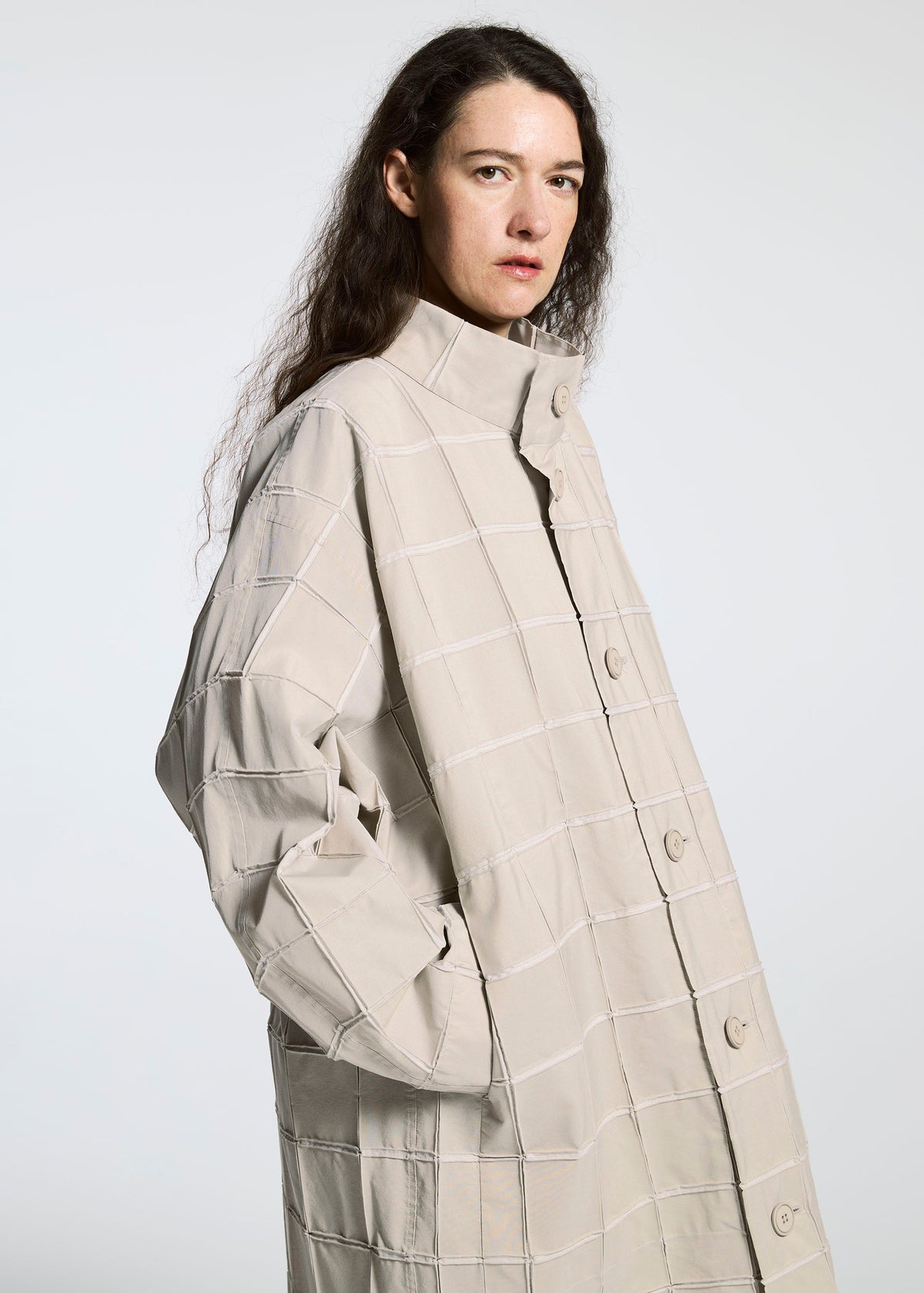 A person with long, wavy brown hair poses against a light grey background, wearing the TYPE-O 002 coat by A-POC ABLE ISSEY MIYAKE—a light beige grid-patterned piece with a high collar made from lightweight stretch material. view 10 of 10
